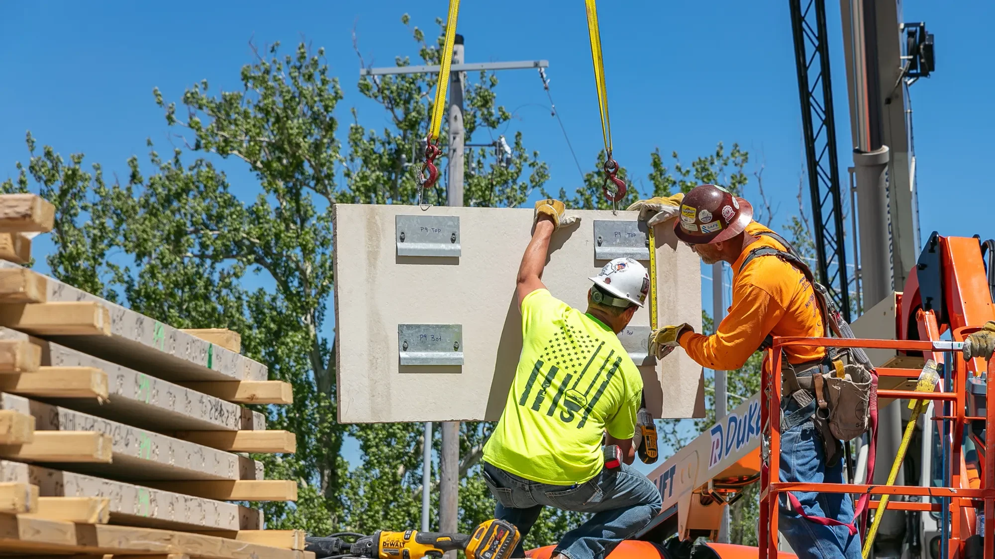 Construction workers installing a large concrete panel on a building site with lumber stacks in the foreground and trees in the background.