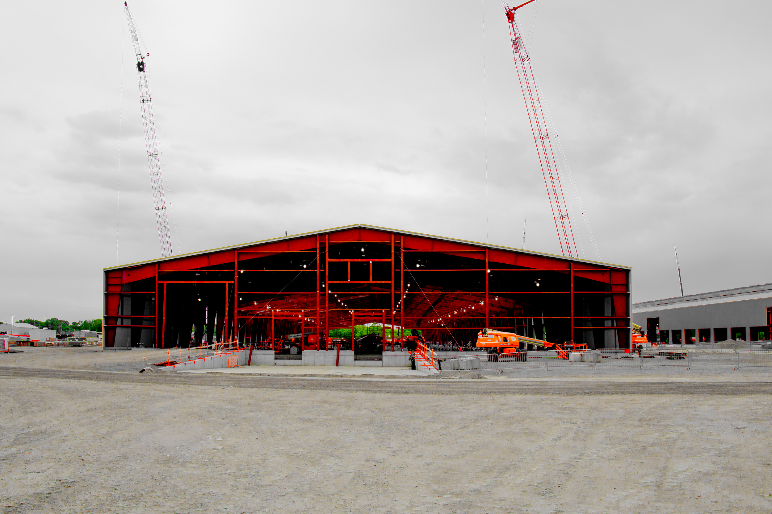 Large red warehouse with overhead lighting, surrounded by construction equipment and fencing, under a cloudy sky.
