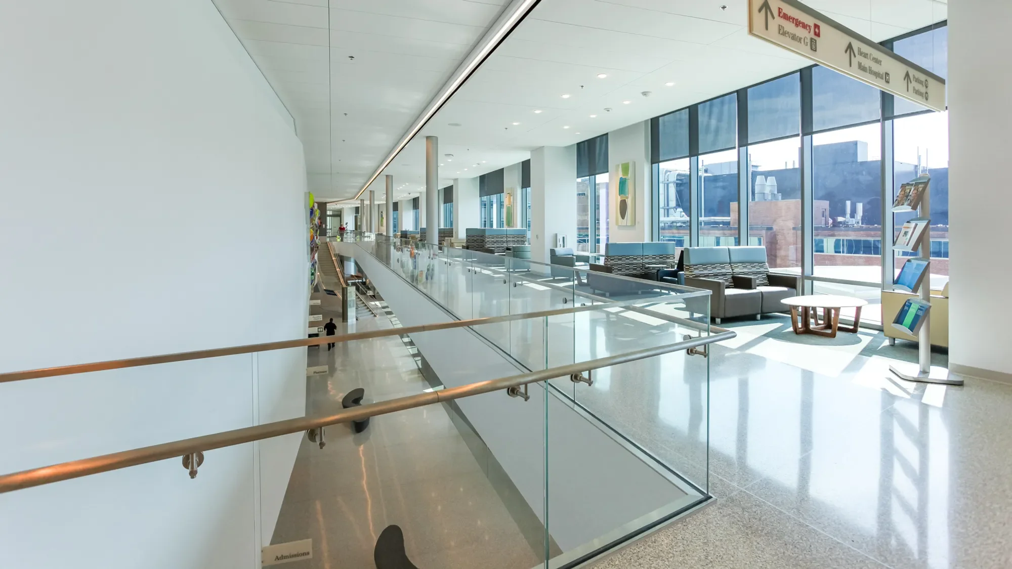 Interior of a modern hospital lounge area with large windows, seating, artwork on the walls, and directional signage overhead indicating emergency, elevator, wing center, main hospital, and parking.