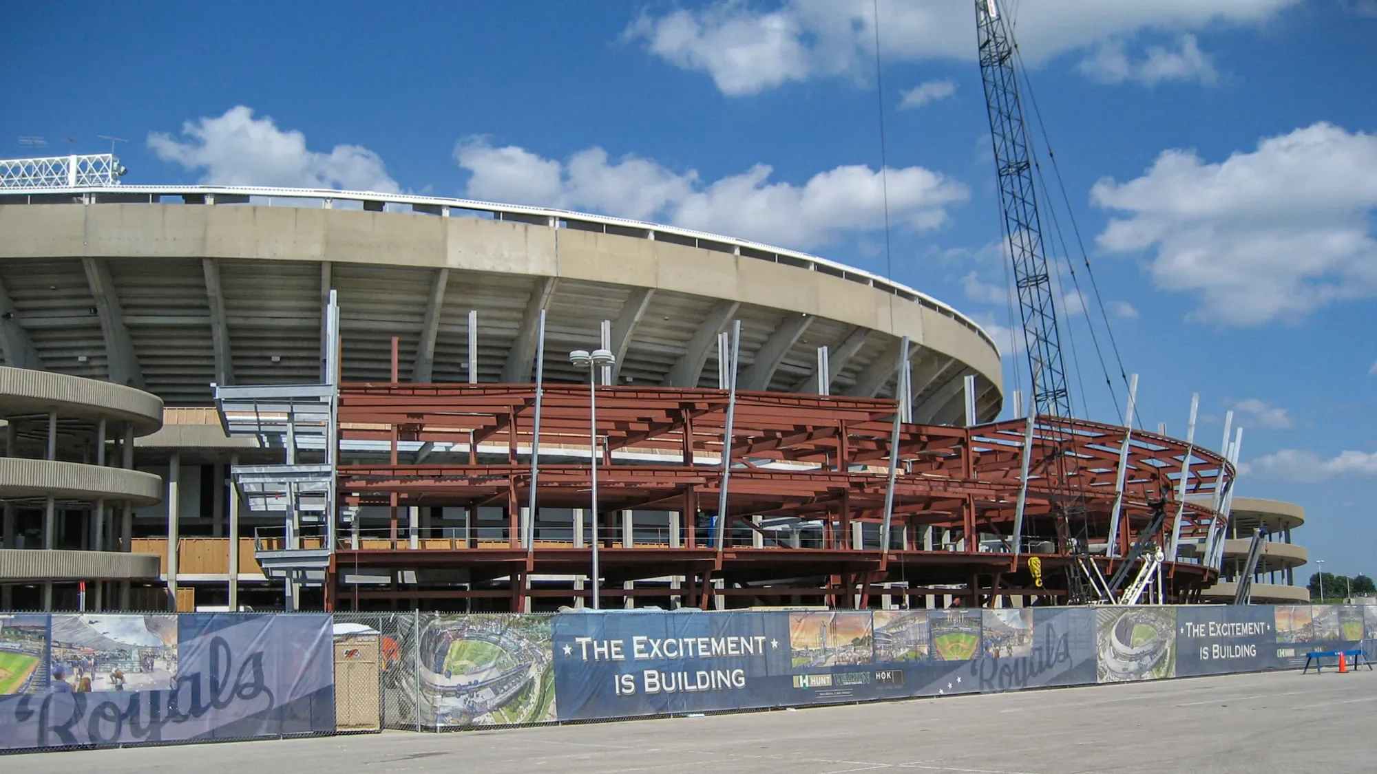 Under construction sports stadium with concrete and steel framework, cranes, and a sign that reads "The Excitement is Building.".