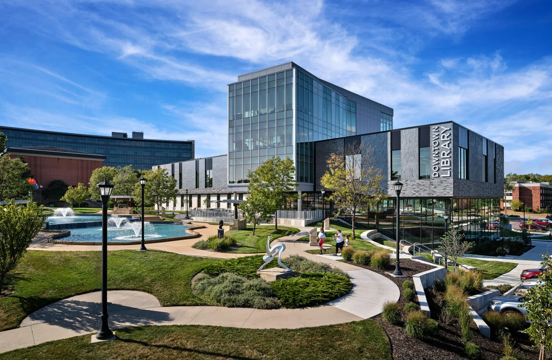 Modern downtown library building with glass and stone facade, surrounded by a landscaped park with trees, benches, fountain, and walking paths under a partly cloudy blue sky.