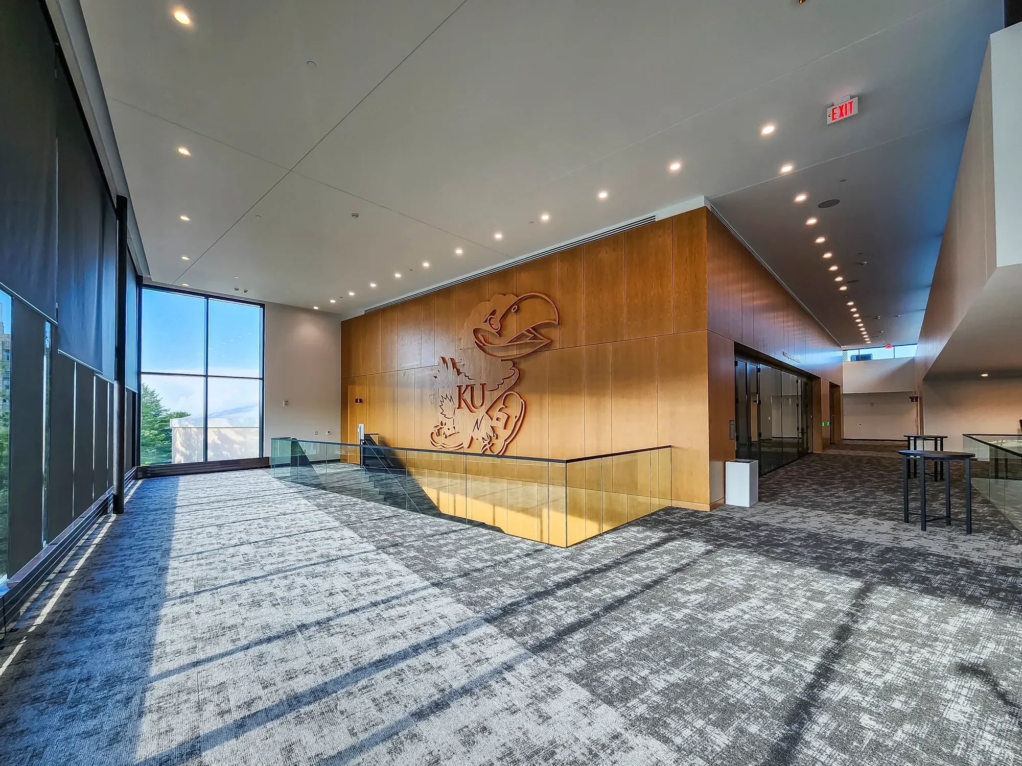 Interior view of a modern lobby area with large windows, wood-paneled wall with a university logo, and patterned carpet. Ceiling has recessed lighting and an exit sign.