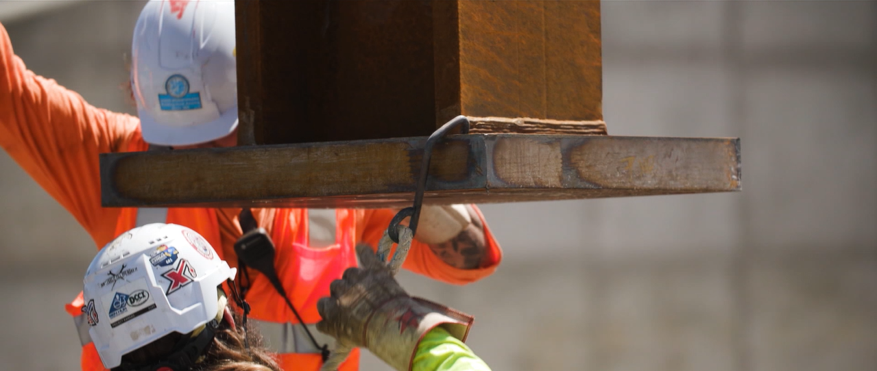 Two construction workers wearing helmets and safety gear lifting a large wooden beam with a hook and chain.