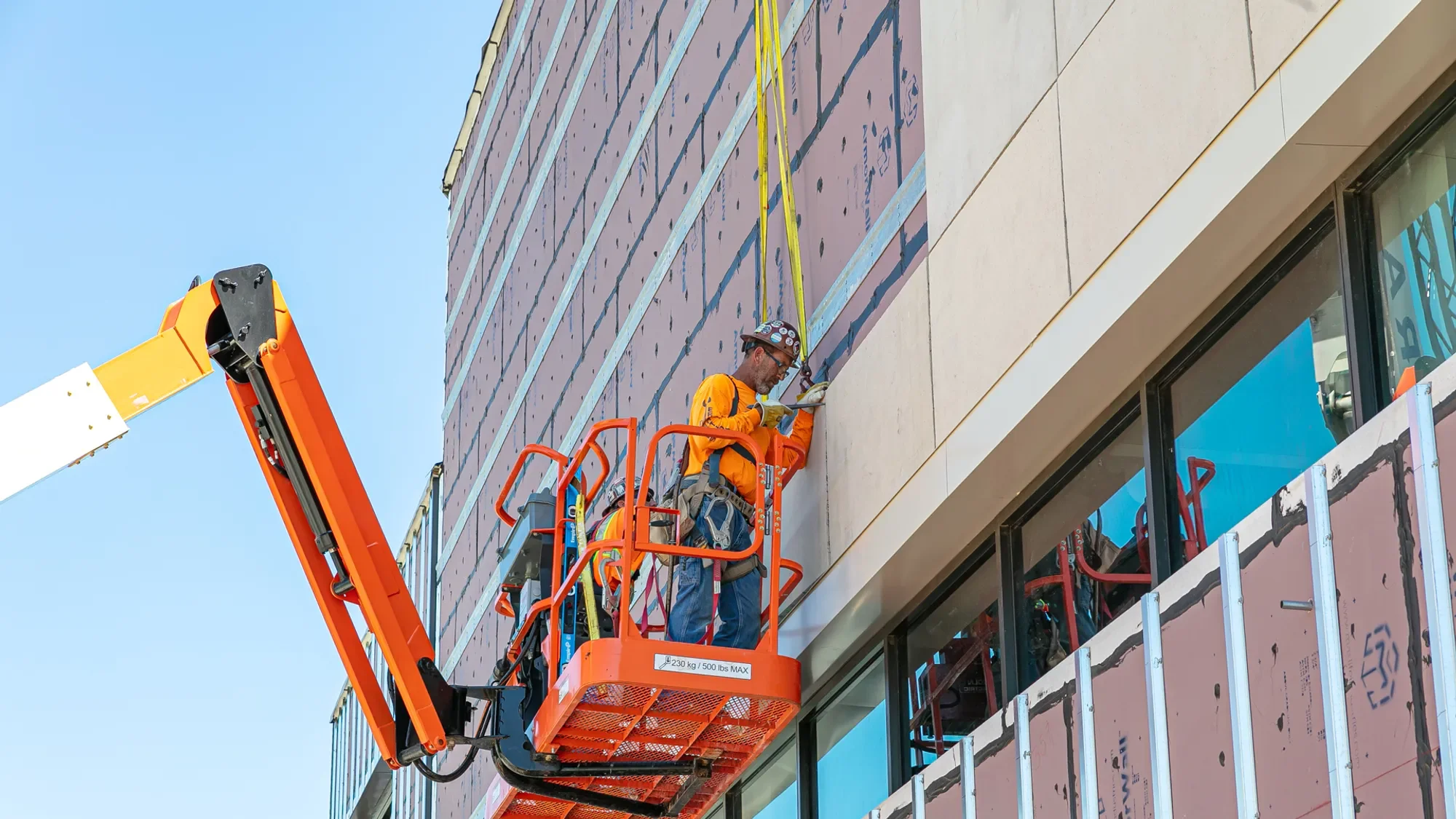 A construction worker in orange safety gear and a helmet is working on the exterior of a building from an elevated lift. The building's facade is partially covered with insulation and structural elements, and a large glass window is visible at the bottom right.