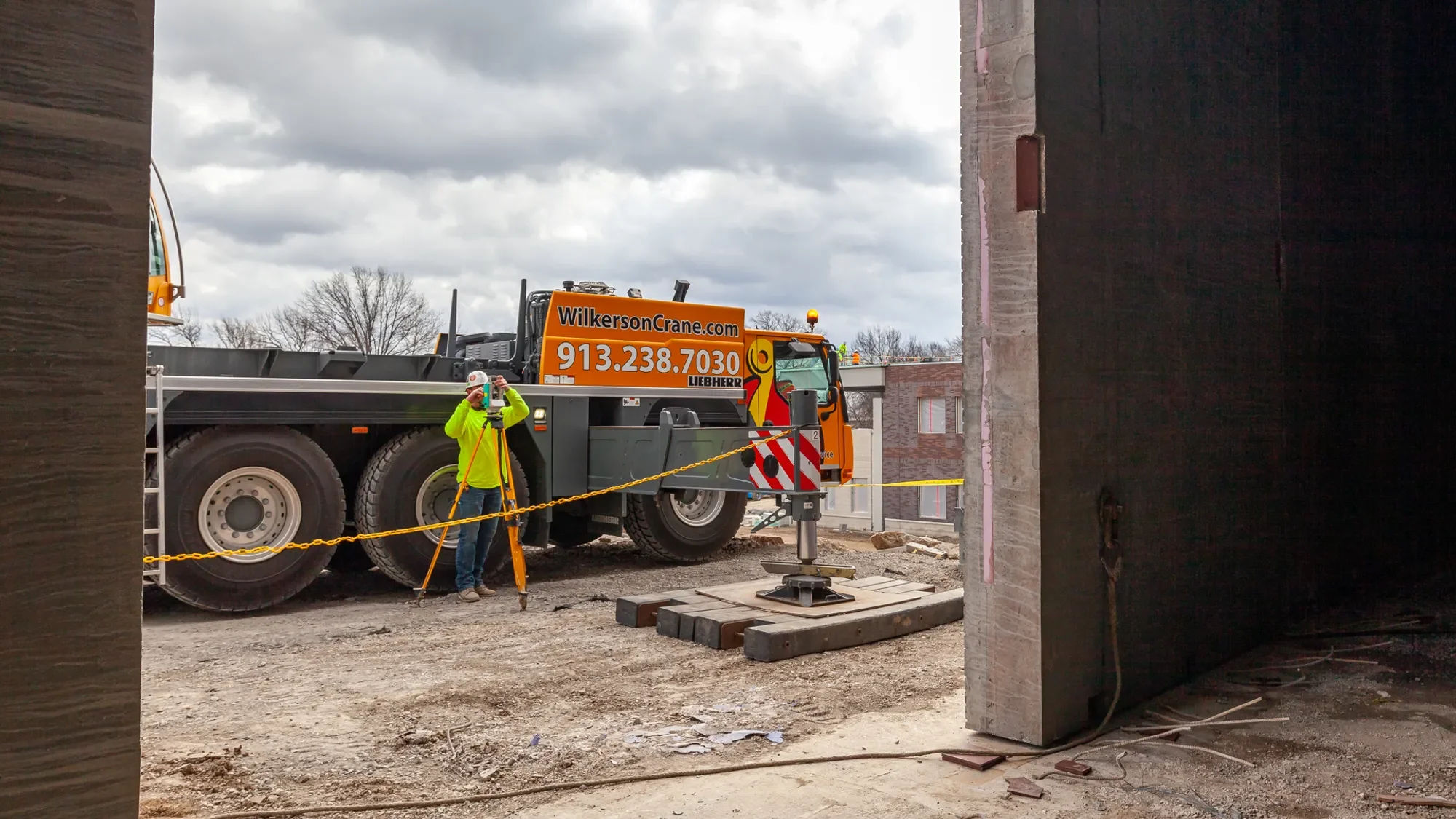 Construction site with a large orange crane truck parked on dirt. A worker in a neon yellow jacket and white hard hat is using surveying equipment, with gray buildings and cloudy sky in the background.