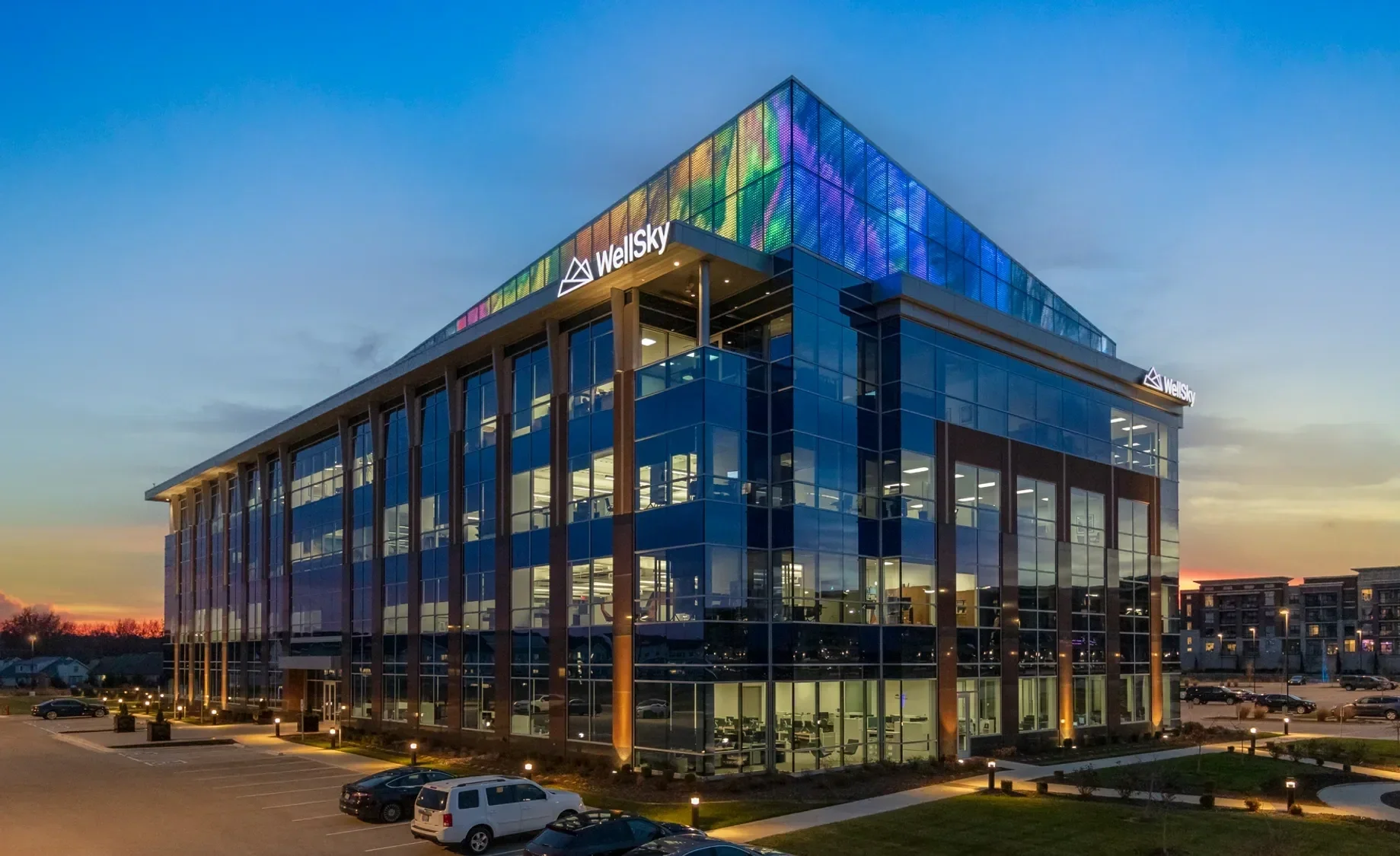 Modern office building with glass exterior illuminated at sunset, displaying the WellSky logo, with parked cars and a landscaped parking lot in the foreground.