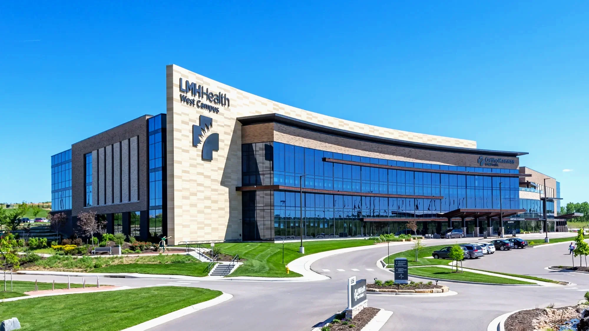 Modern medical building with glass windows and a sign that reads 'LMH Health West Campus'. Parking lot in front with cars, landscaped greenery, and clear blue sky.