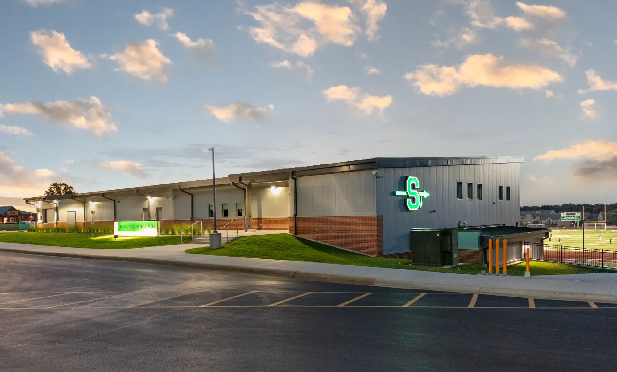 Exterior of a school or community center building during sunset, with a green and white illuminated logo on the side, surrounded by a parking lot and a sports field in the background.