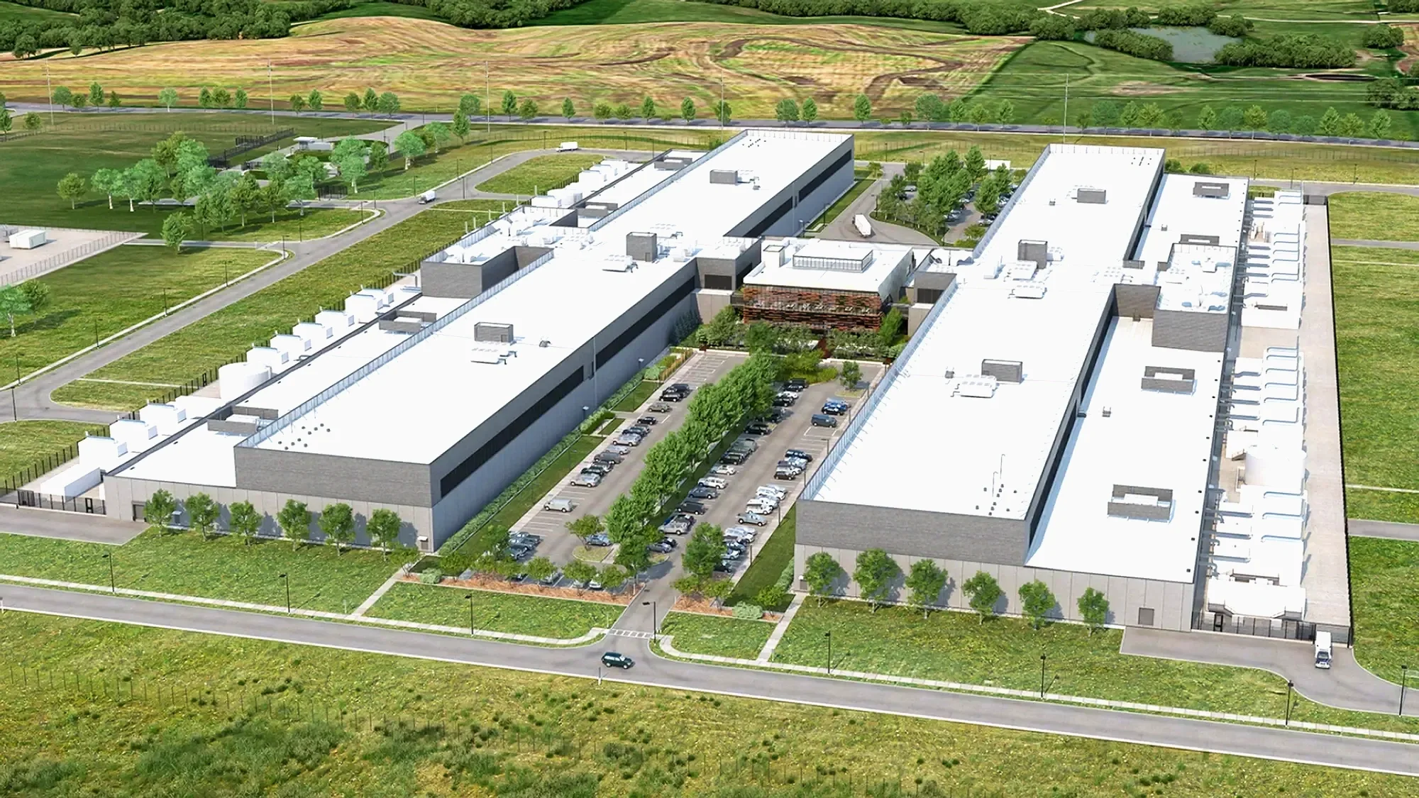 Aerial view of a large modern industrial or commercial building with a white roof, surrounded by green grass, trees, parking lots with cars, and a landscape of hills and fields in the background.