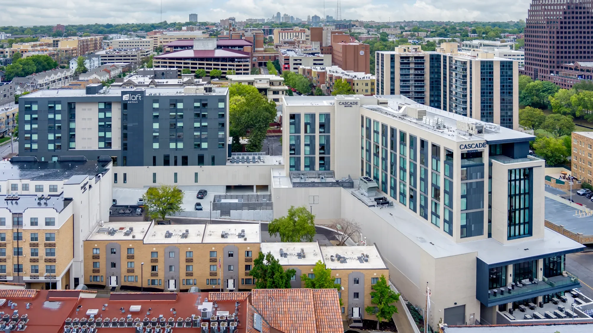 Aerial view of a city skyline featuring various high-rise buildings, including the Aloft and Cascade Hotels, as well as residential and commercial structures, streets, and green trees.