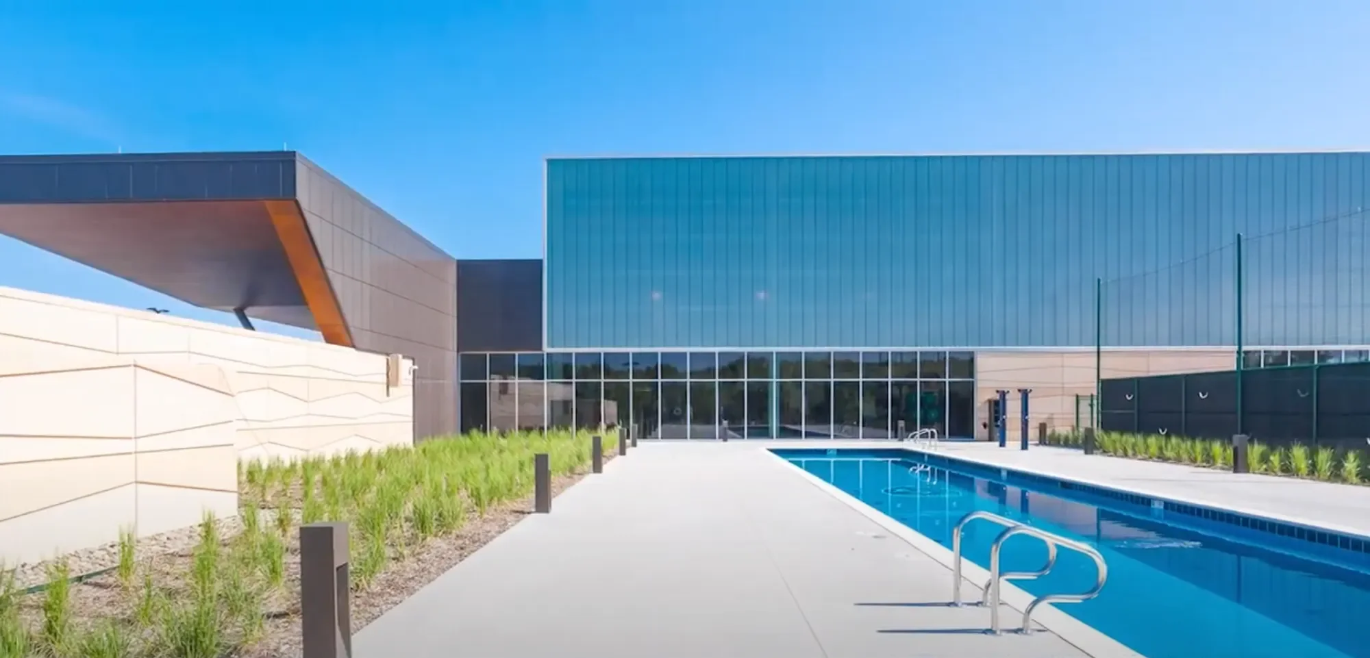 Modern building with large glass windows, an outdoor swimming pool, and landscaped plants under a clear blue sky.