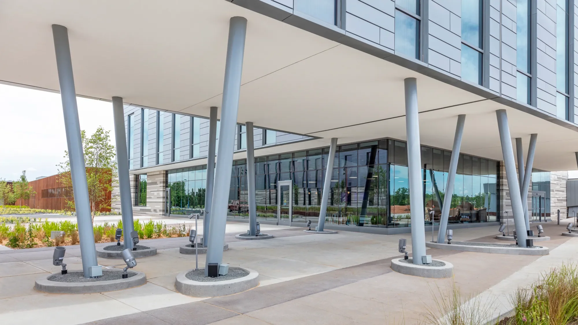 Modern building with glass windows and an overhang supported by angled columns. In the foreground, there are landscape and outdoor lighting fixtures.