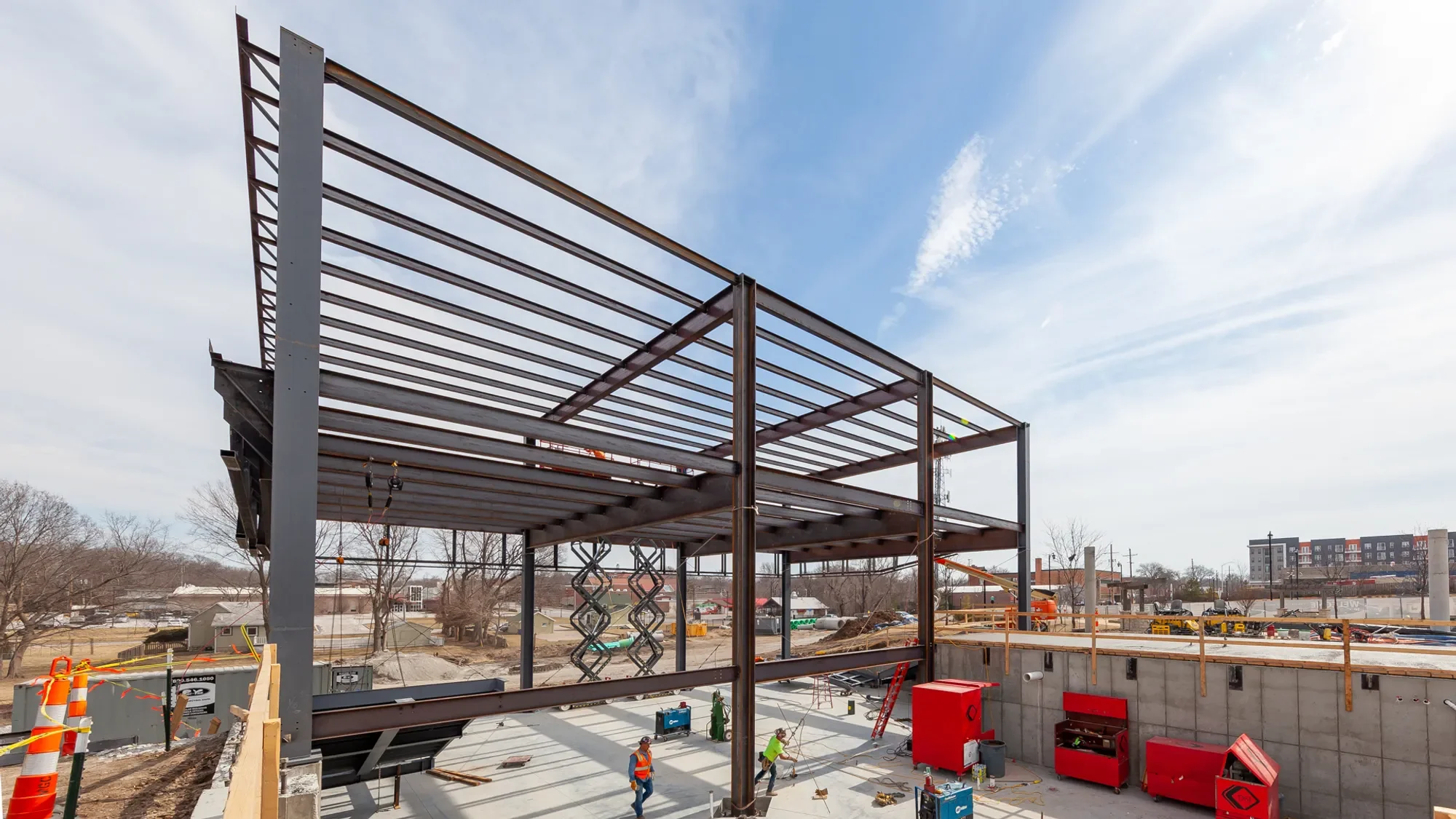 A construction site with a steel framework for a building, workers in safety vests, and construction equipment under a partly cloudy sky.