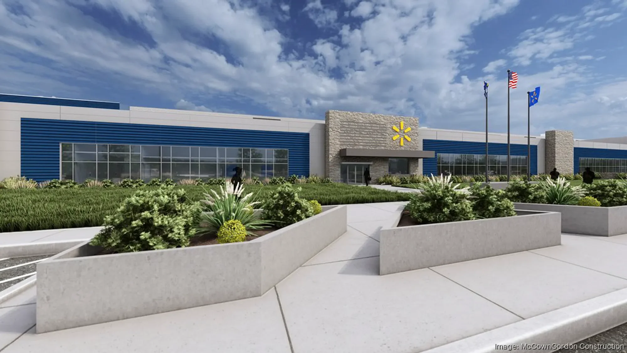 A modern commercial building with blue and white exterior, large glass windows, and a Walmart logo near the entrance. There are three flagpoles with American, corporate, and state flags, and landscaped greenery with flowering plants in the foreground. The sky is partly cloudy.