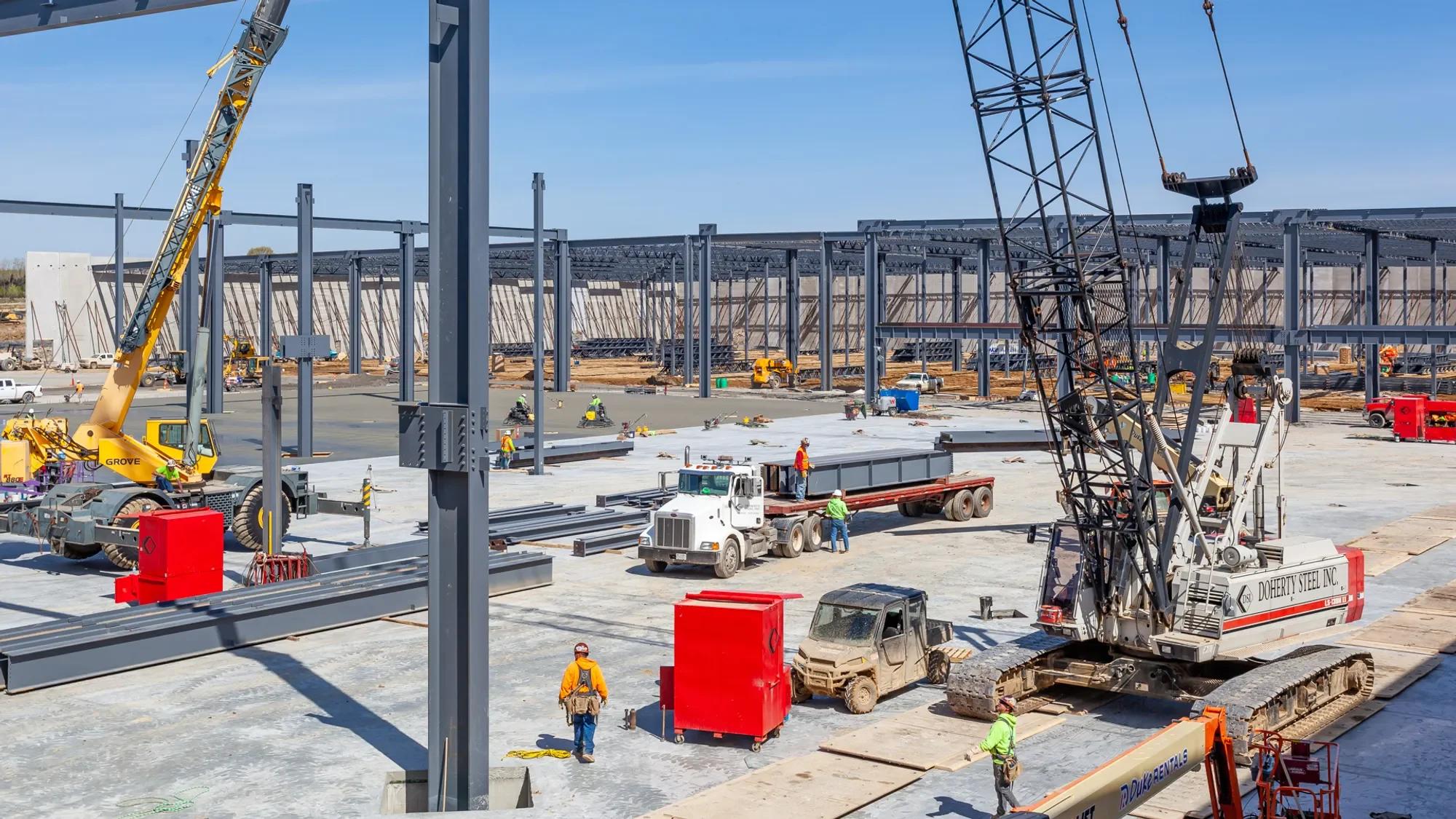 Construction site with workers, cranes, metal framework, and trucks working on building structure under blue sky.