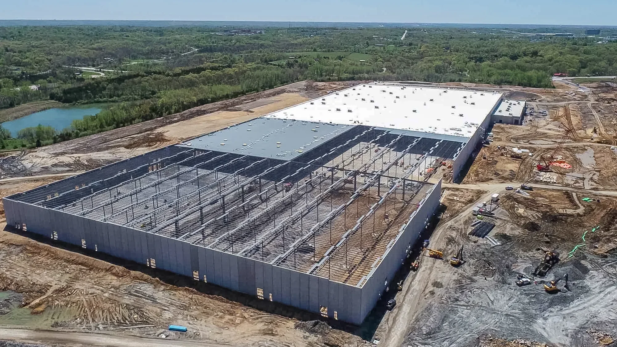 Construction site with a large building under construction, featuring a metal framework and partially installed roof panels. Surrounding area has dirt and construction equipment, with a scenic background of green trees, a pond, and distant roads.