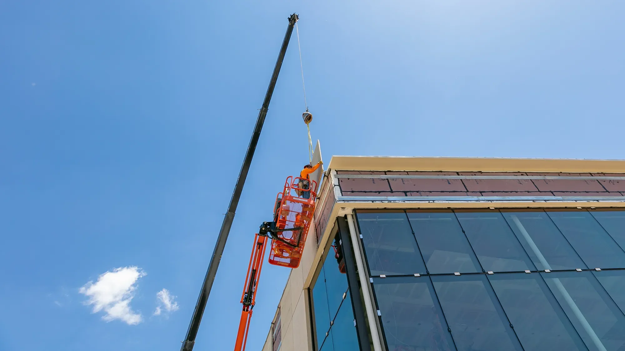 A construction worker in an orange shirt working on the exterior of a modern building with large glass windows, using a crane with a basket lift to reach the upper part of the building against a blue sky with some clouds.