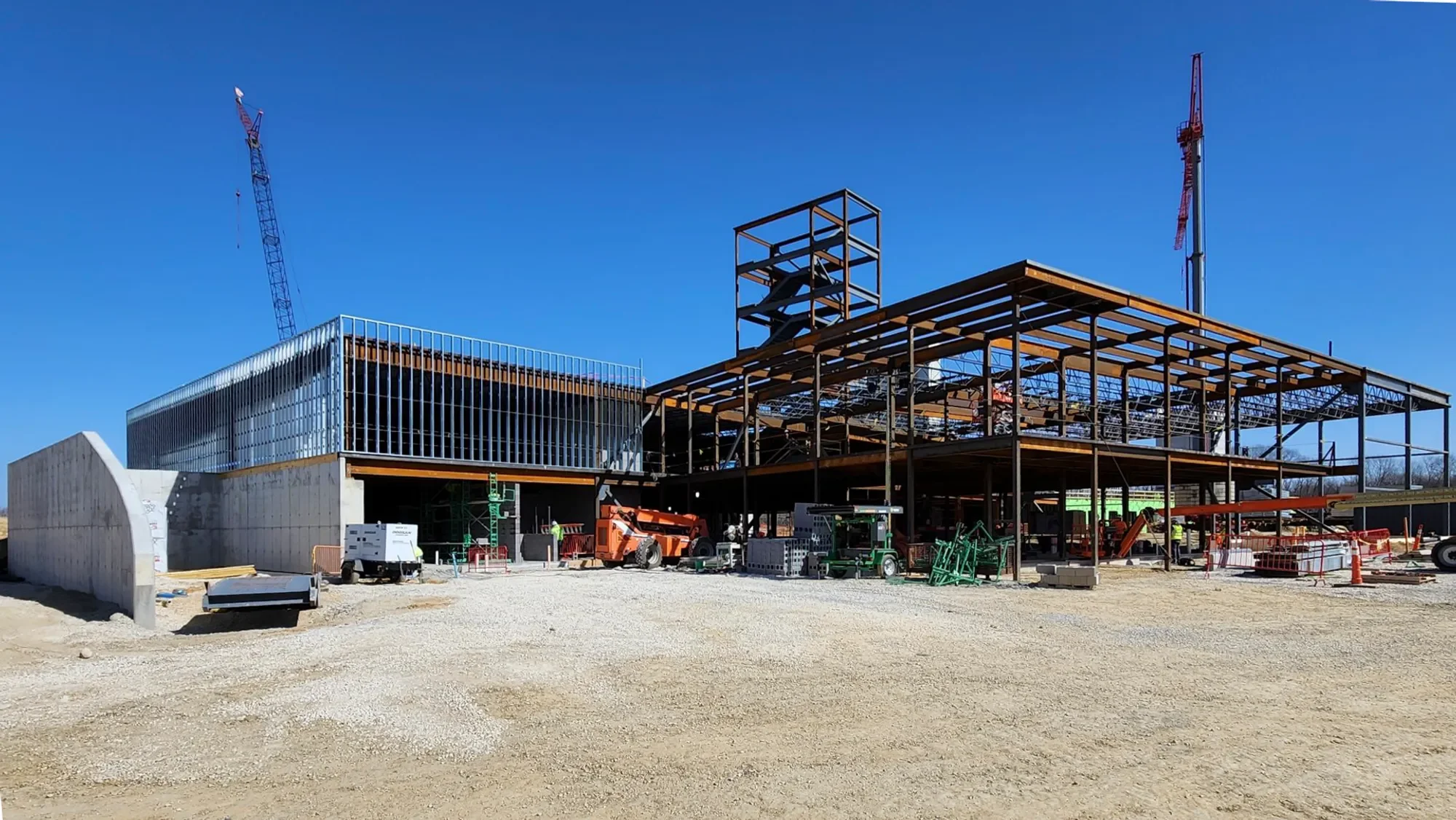 A construction site with a partially built multi-story structure, cranes, and construction equipment under a clear blue sky.