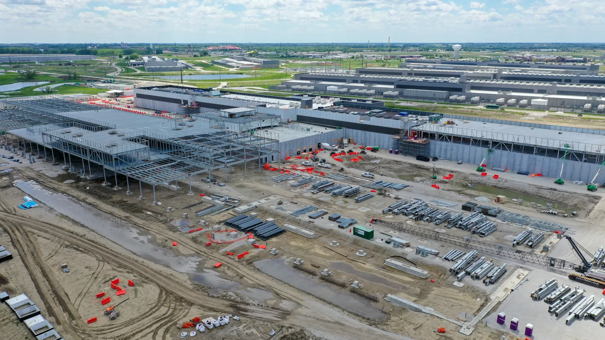 Aerial view of a large industrial building under construction, with construction materials, equipment, and cranes visible on site, and solar panels installed on the roof.