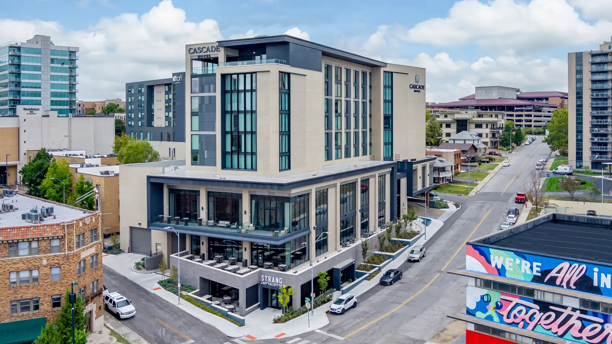 Aerial view of modern multi-story hotel building named Cascade Hotel, with surrounding high-rise buildings, streets, and parked cars in an urban area.