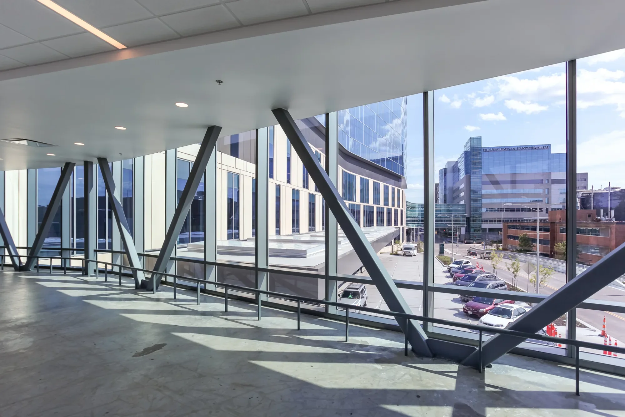 Interior view of a modern building with large glass windows showing a cityscape outside, including tall office buildings and parking lot. The ceiling has recessed lighting and the floor is polished concrete, with structural steel beams supporting the area.