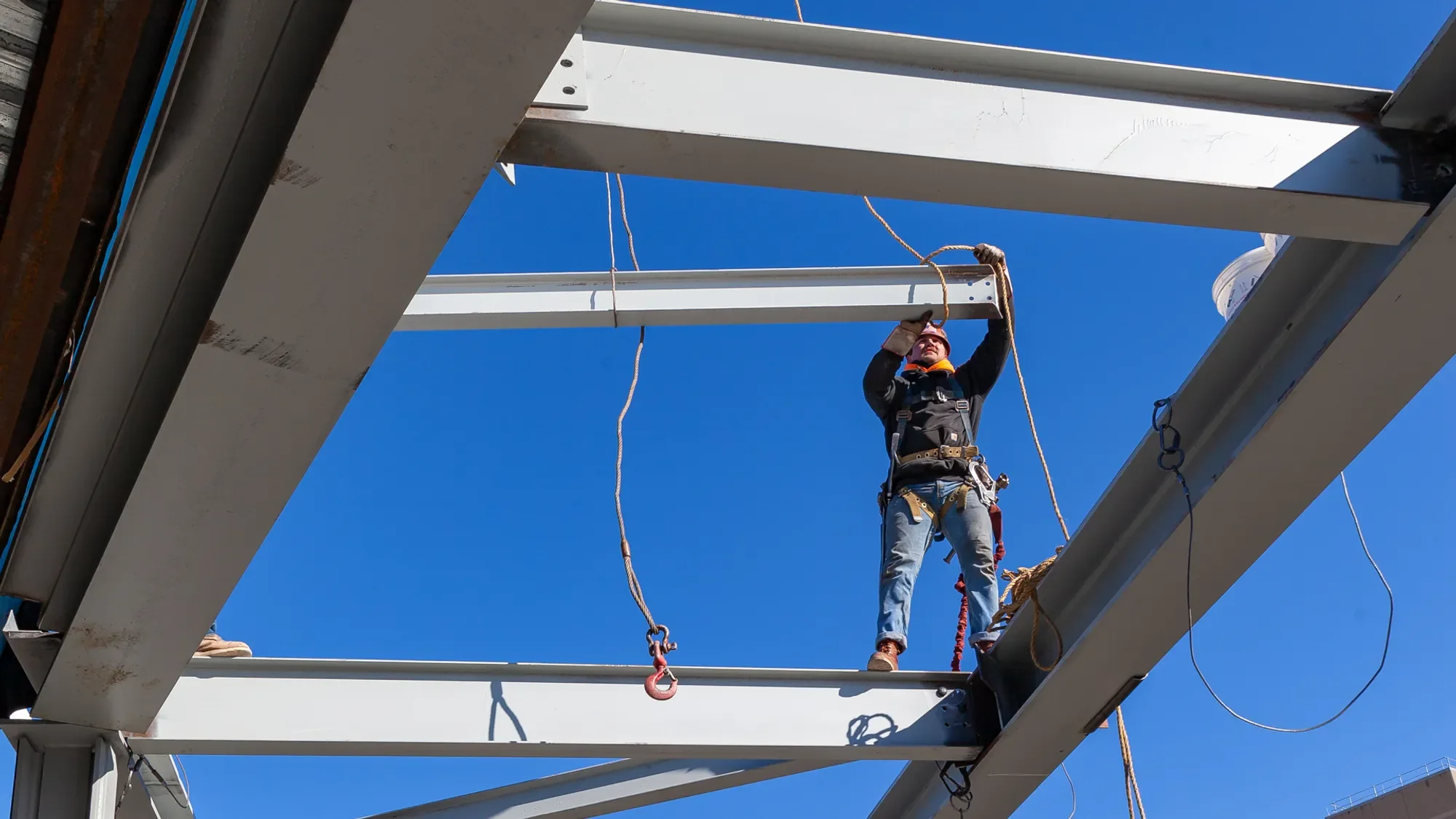 A construction worker wearing safety gear is working on steel beams at a construction site under a clear blue sky.