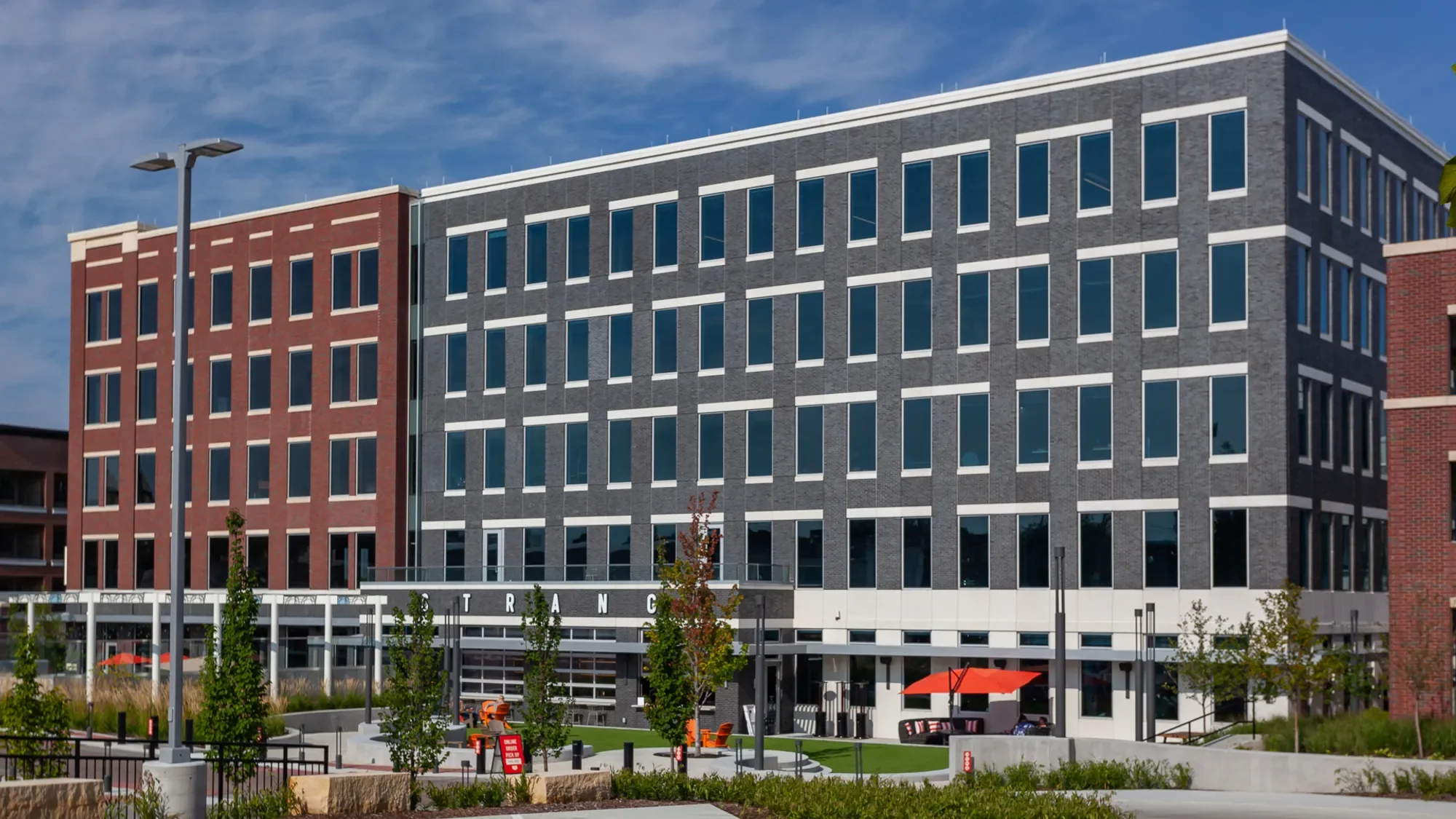 Modern multi-story office building with red and gray brick facade, numerous windows, outdoor seating area, trees, and orange umbrellas, under a partly cloudy sky.