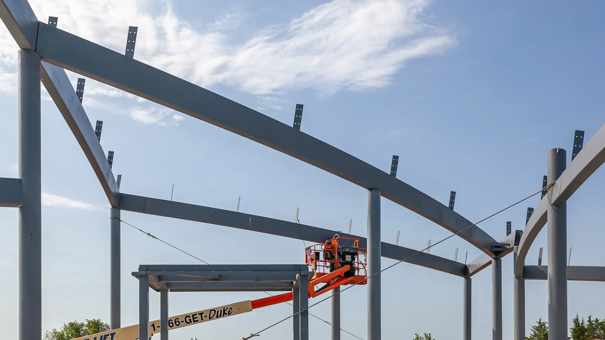Construction workers using a lift to build a large steel frame structure under a partly cloudy sky.