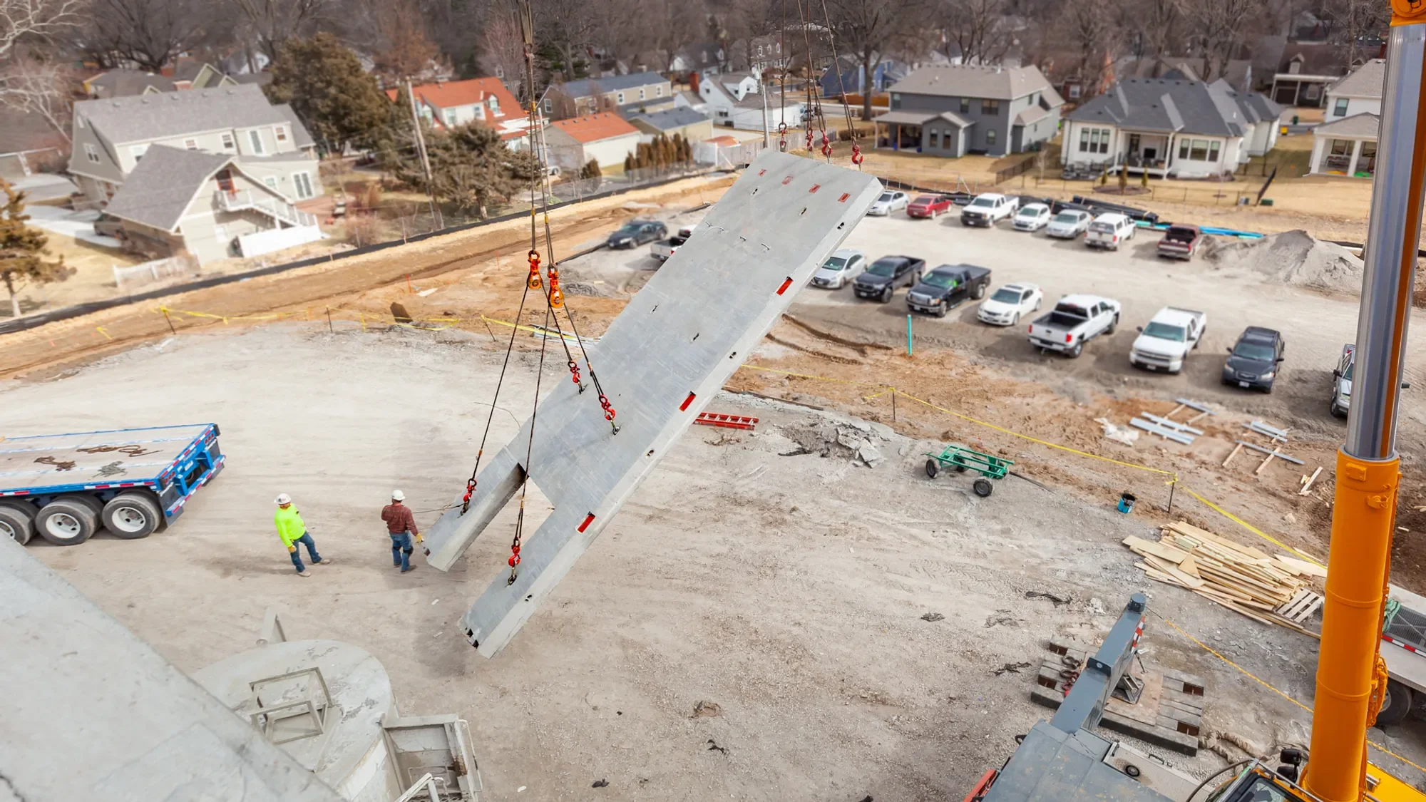 Construction workers are installing a large concrete panel at a building site, with a crane lifting the panel into place. The site has a gravel surface, construction equipment, and parked cars, with residential houses in the background.