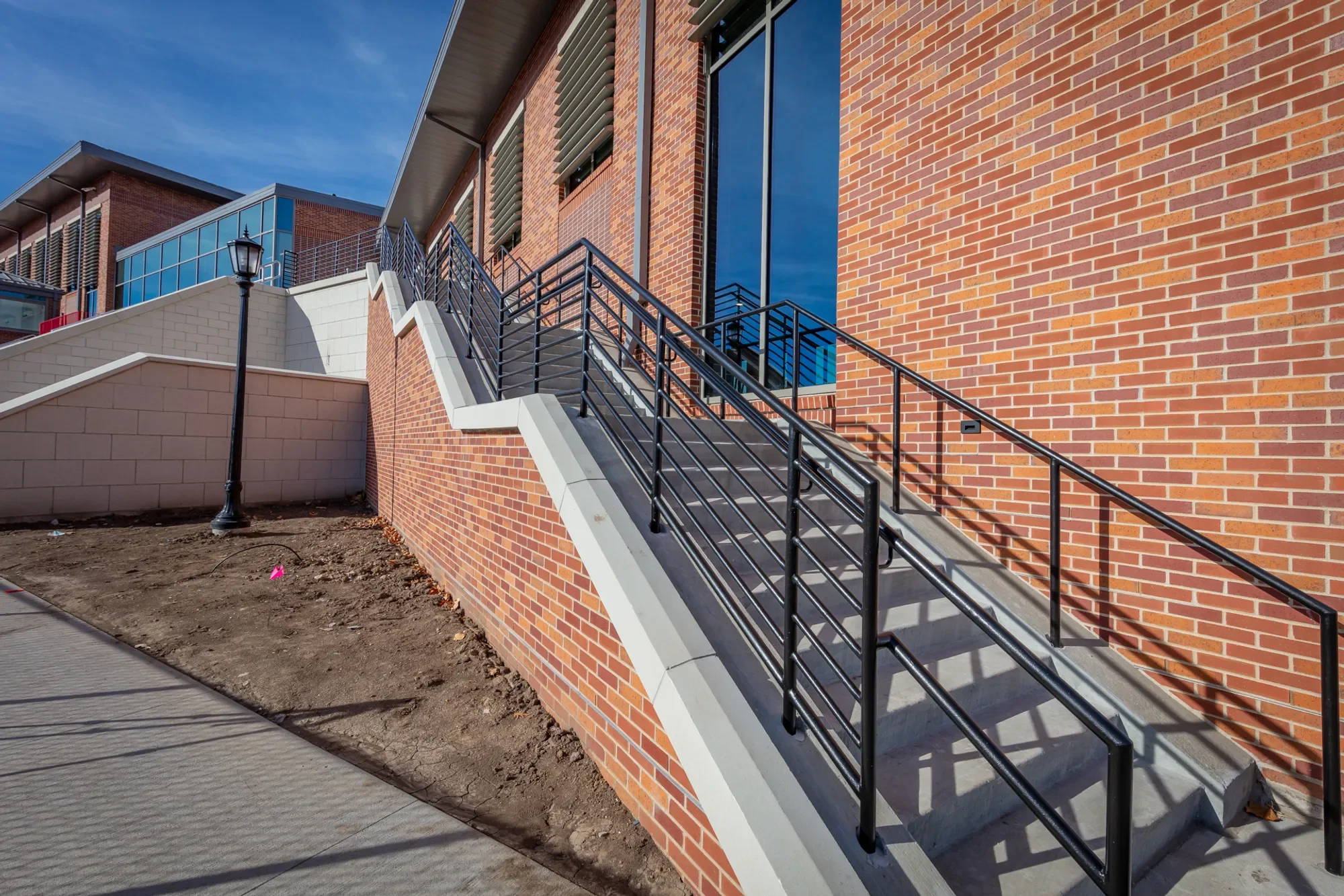A modern building exterior made of red brick with large glass windows and metal railings on a staircase leading up to an entrance. The image shows an outdoor scene with a clear blue sky and part of a sidewalk and dirt area in the foreground.