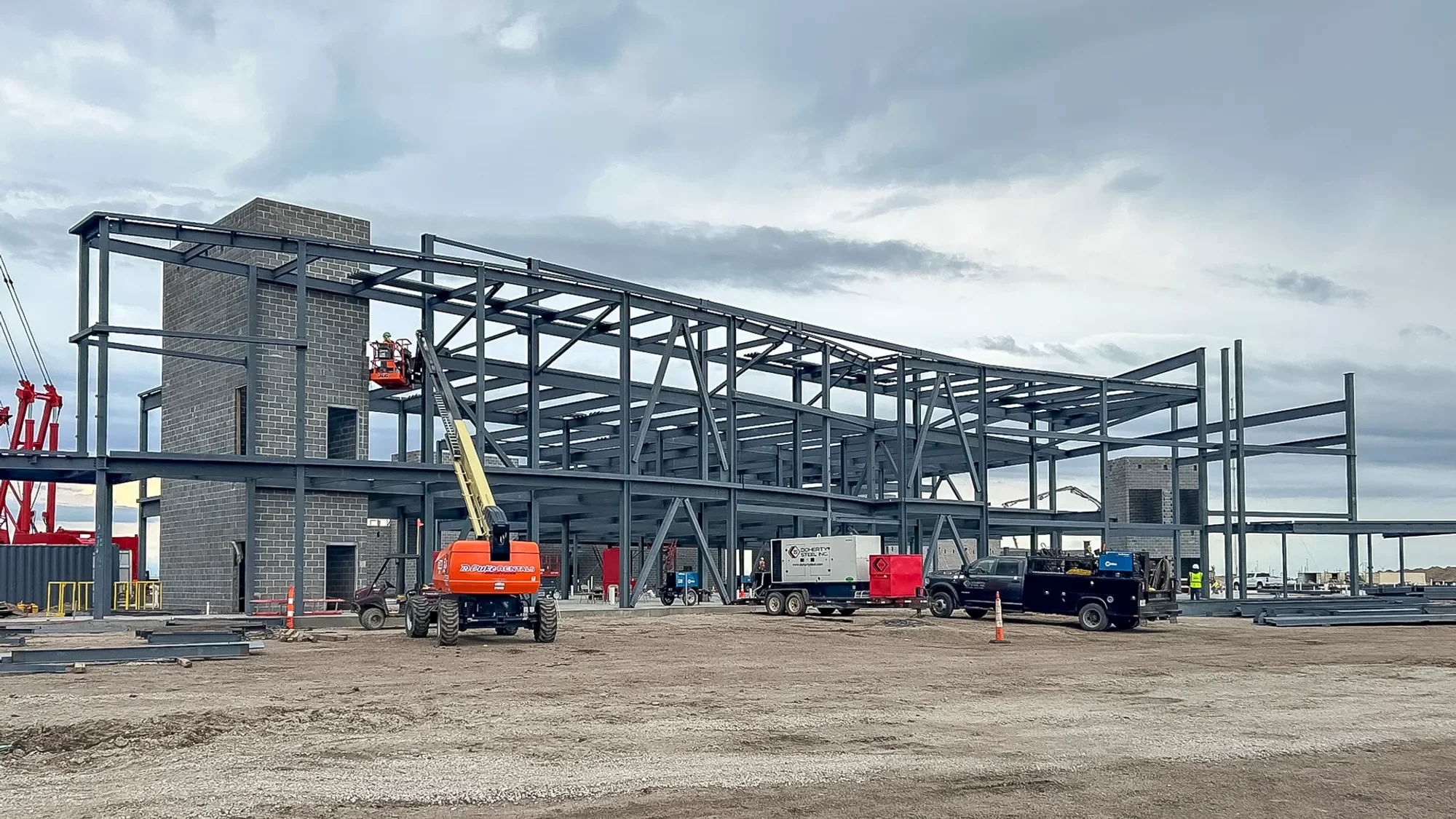 Construction site with steel framework being assembled for a building, several construction vehicles and workers present, cloudy sky overhead.