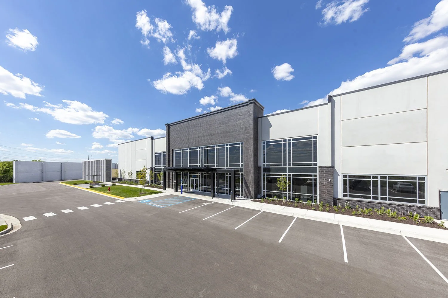 A modern commercial building with large glass windows, a parking lot with marked spaces, some small trees and landscaping, under a bright blue sky with clouds.