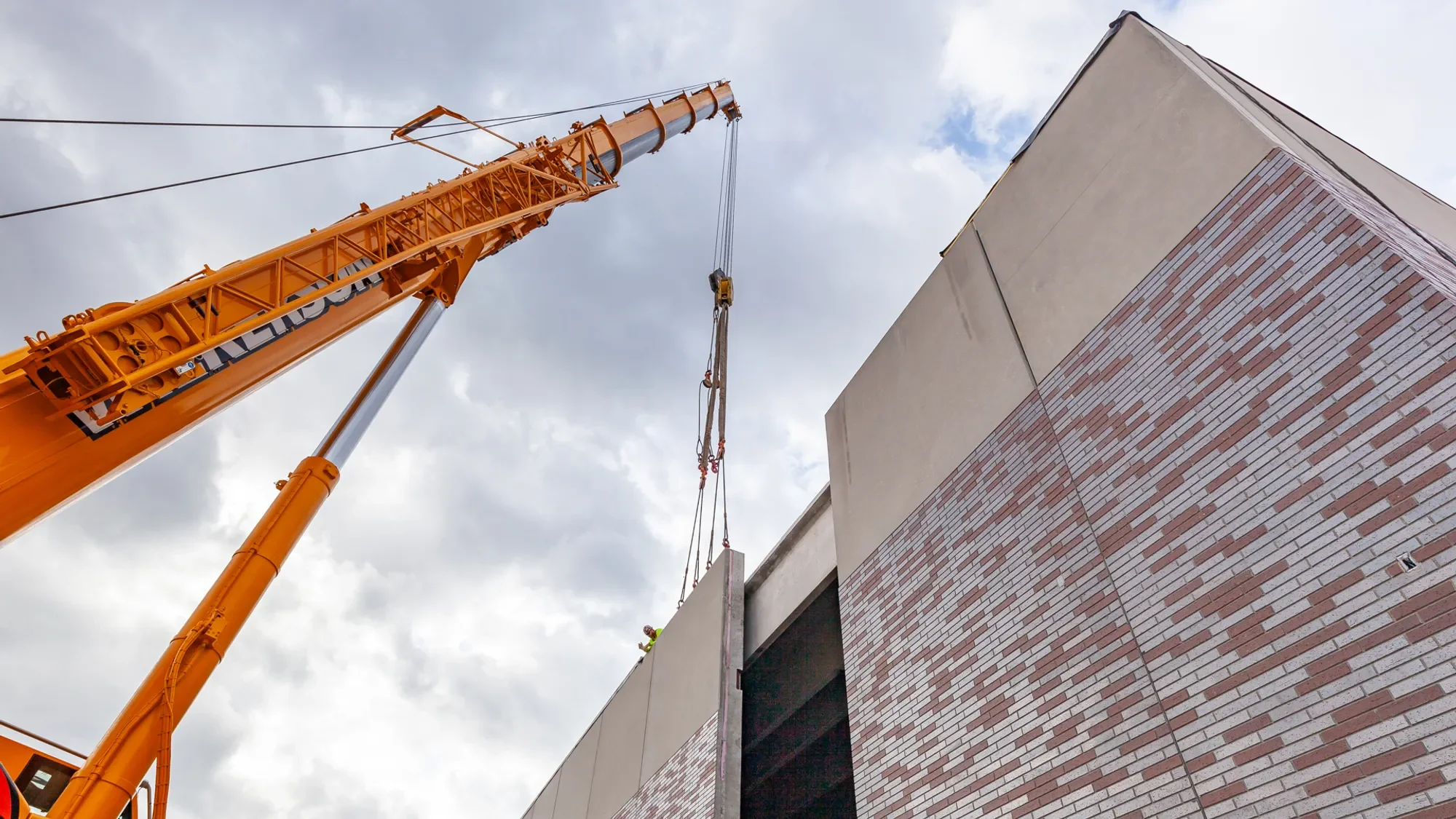 Large orange crane lifting a concrete slab at a construction site with a building featuring brick and beige panels and cloudy sky in the background.