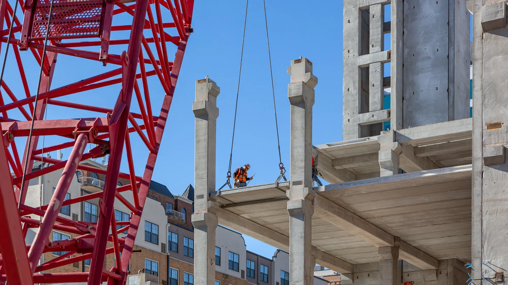 Construction site with a worker wearing orange safety gear, a hard hat, and gloves, standing on the concrete floor of a building under construction. A large red crane is nearby, with parts of its structure visible, and several concrete columns are being installed against a clear blue sky.