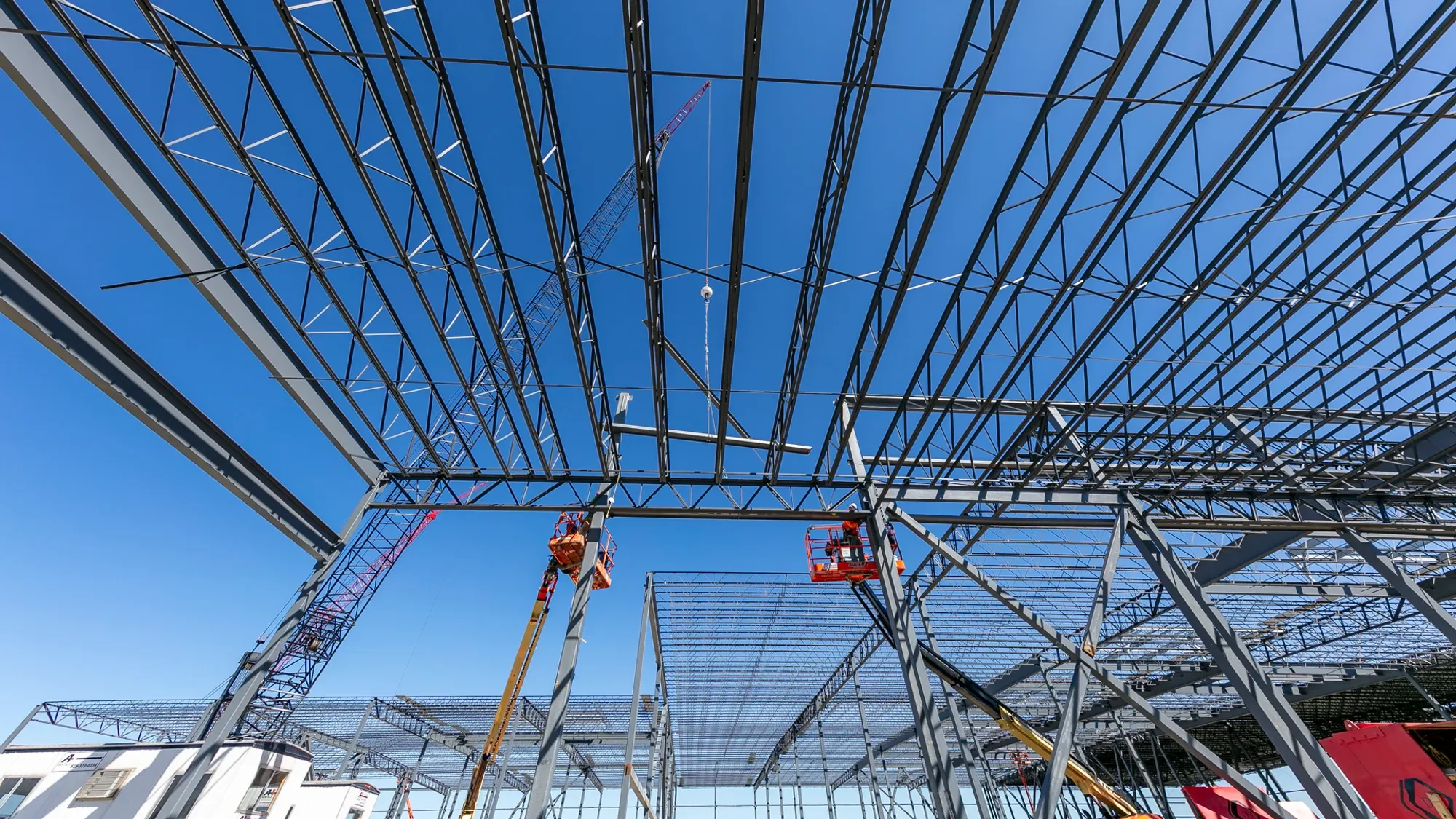 Construction site with a steel framework for a building, two elevated work platforms with workers, cranes, and a clear blue sky.