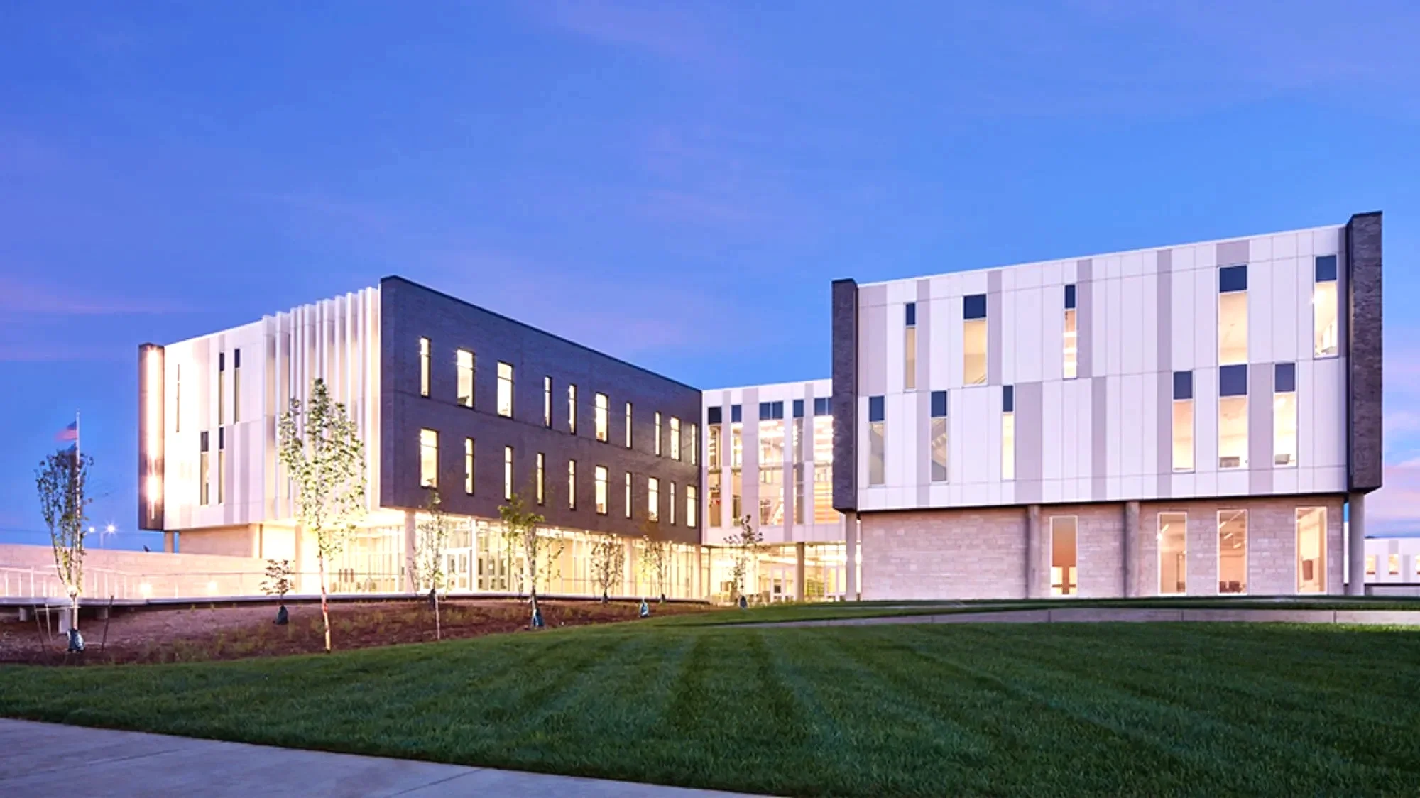 Modern multi-story office building with illuminated windows and a landscaped lawn in the foreground, under a twilight sky.