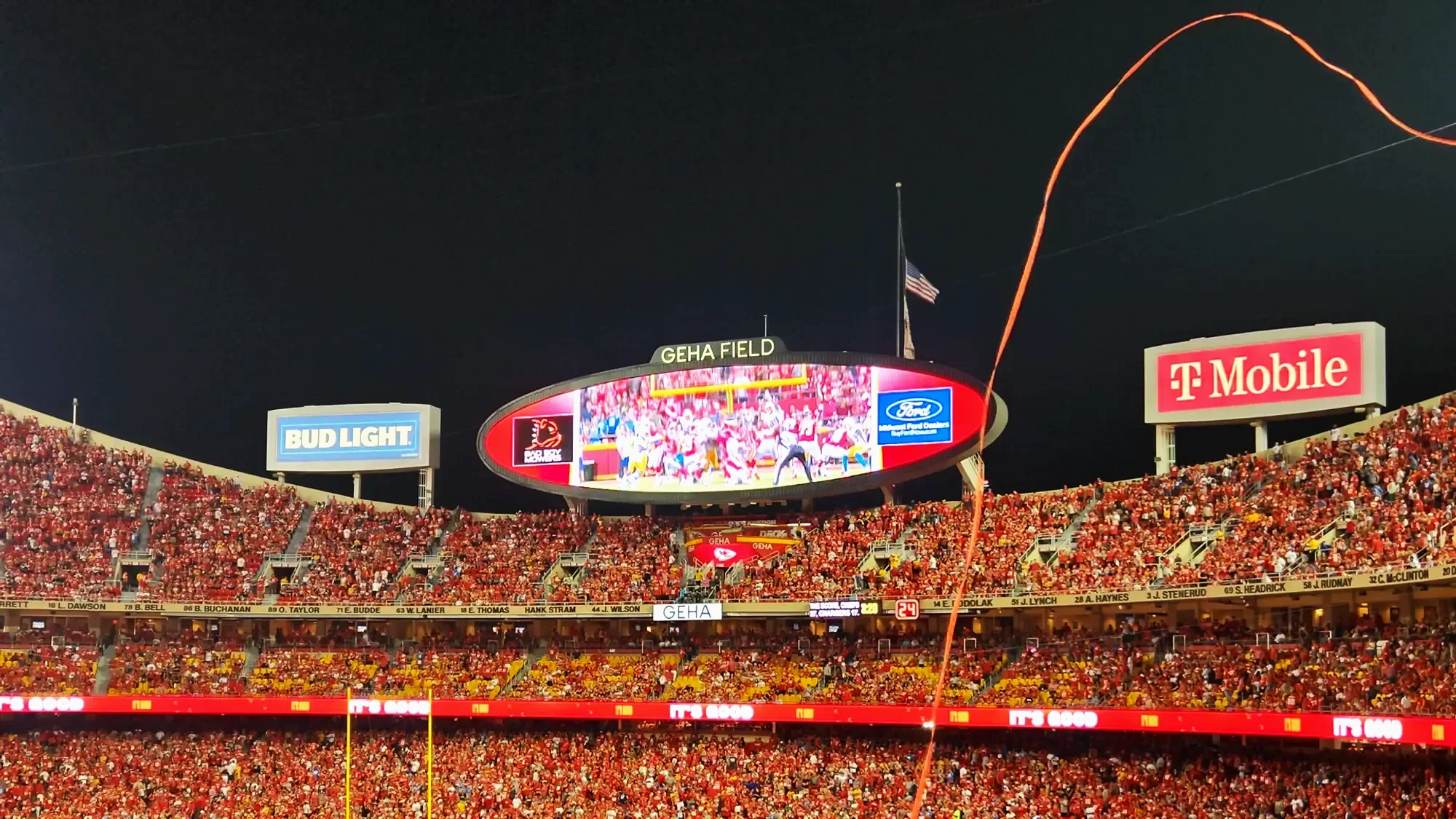 Nighttime view of GEHA Field at a stadium filled with millions of red seats and a large crowd. The stadium's video scoreboard displays a live game with players and cheerleaders, with the flags of Kansas City and the United States flying above. Orange fireworks are visible in the sky.