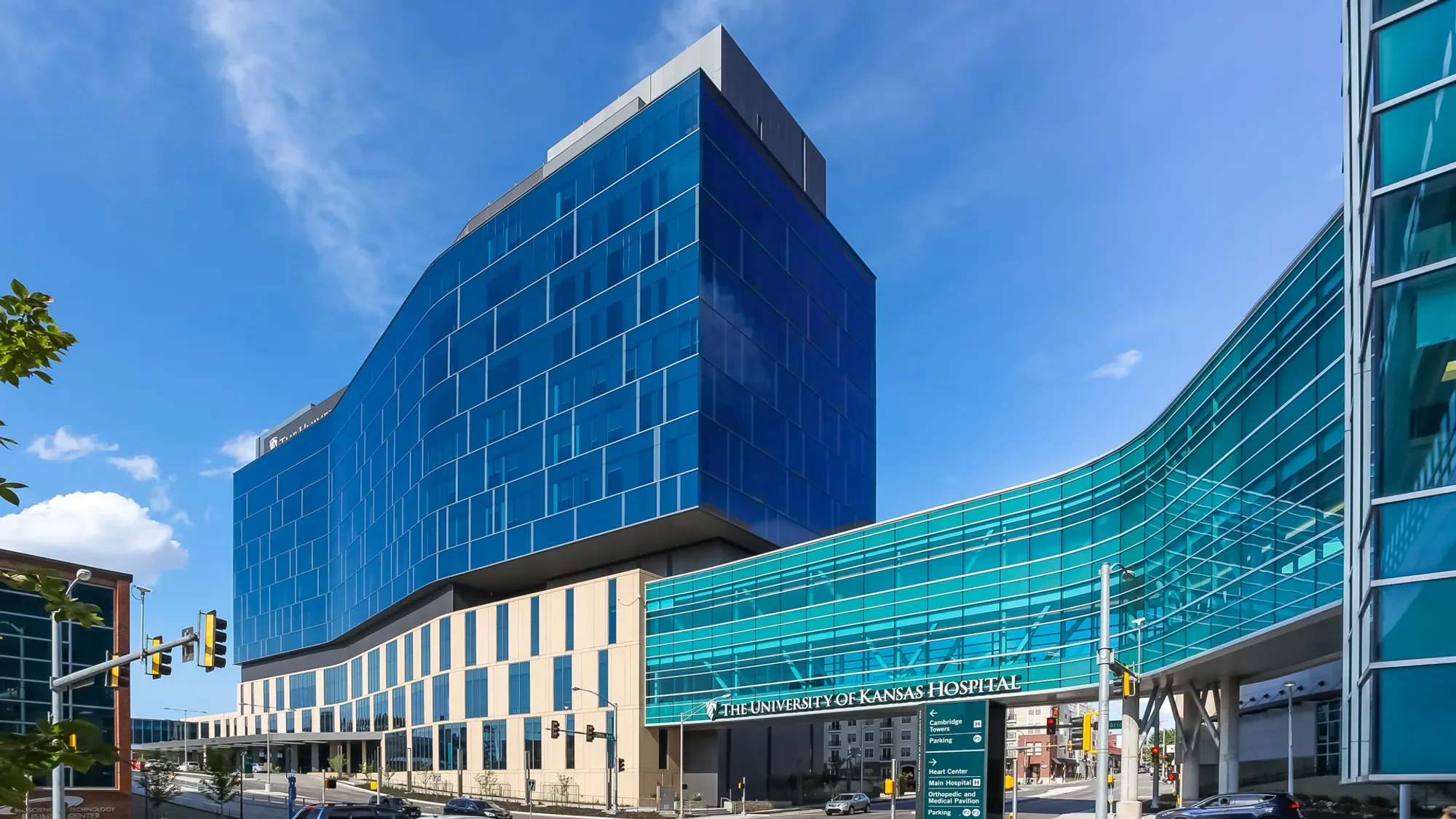 Modern blue glass hospital building with signage reading 'The University of Kansas Hospital,' set against a blue sky with some clouds.