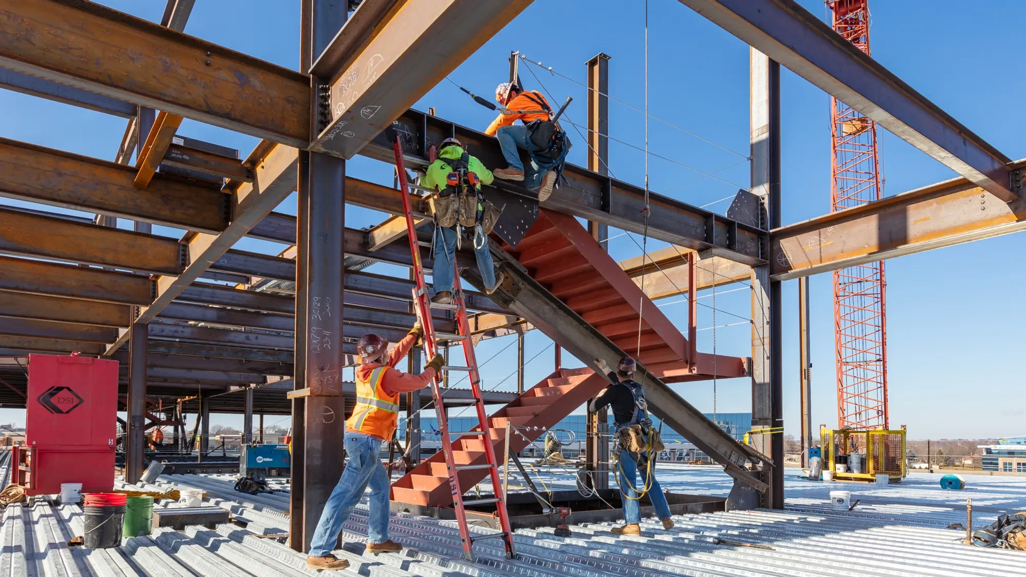 Workers assembling steel framework on a construction site, with a staircase and crane in the background, under a clear blue sky.