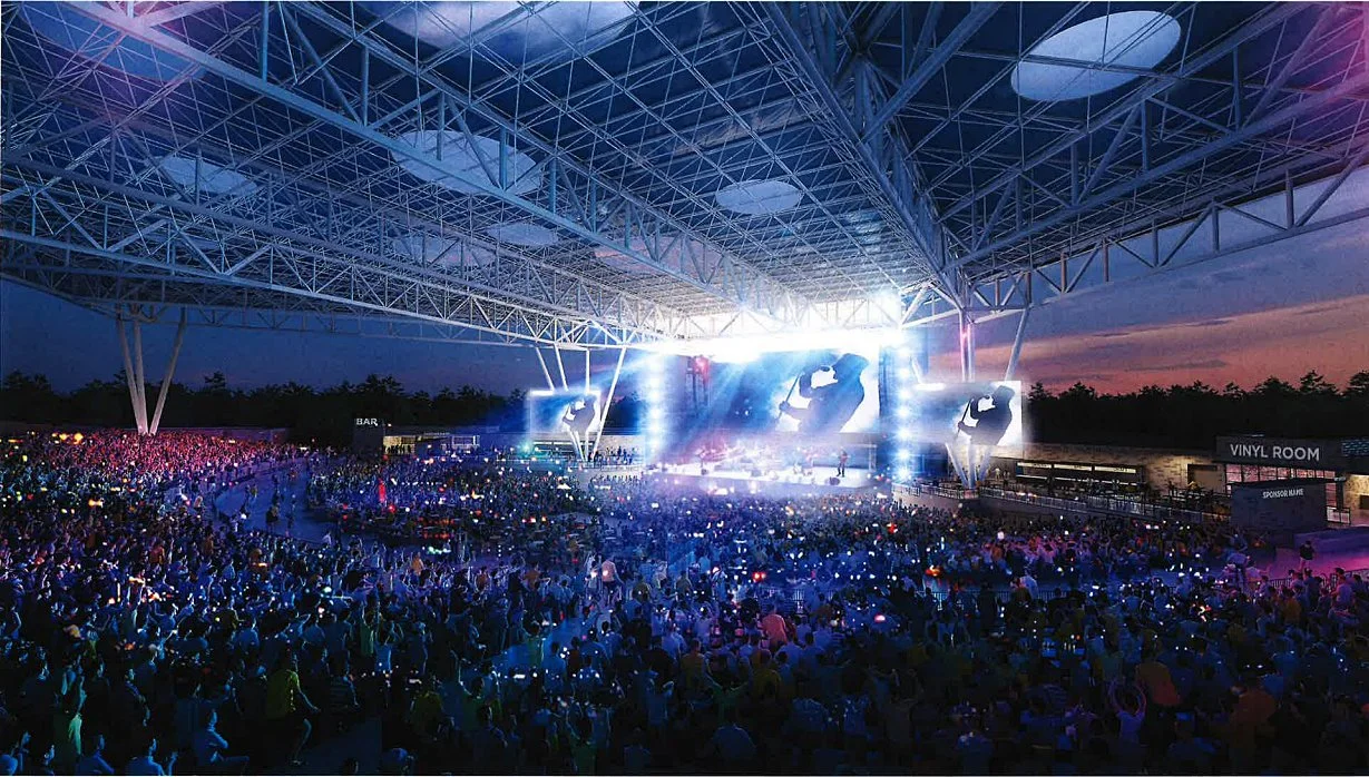 A large outdoor concert with a full crowd, stage lighting, and large screens at dusk or night under an open metal roof structure.