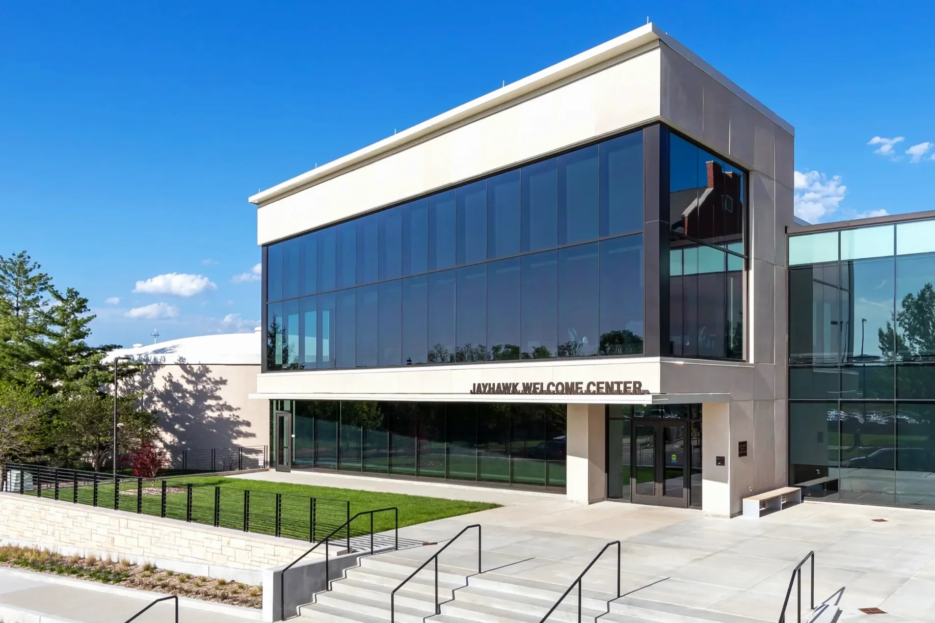 Modern two-story building with glass windows and a white facade, labeled 'JAYHAWK.WELCOME CENTER,' with stairs and a ramp leading to the entrance, surrounded by trees and a well-maintained lawn, under a bright blue sky.