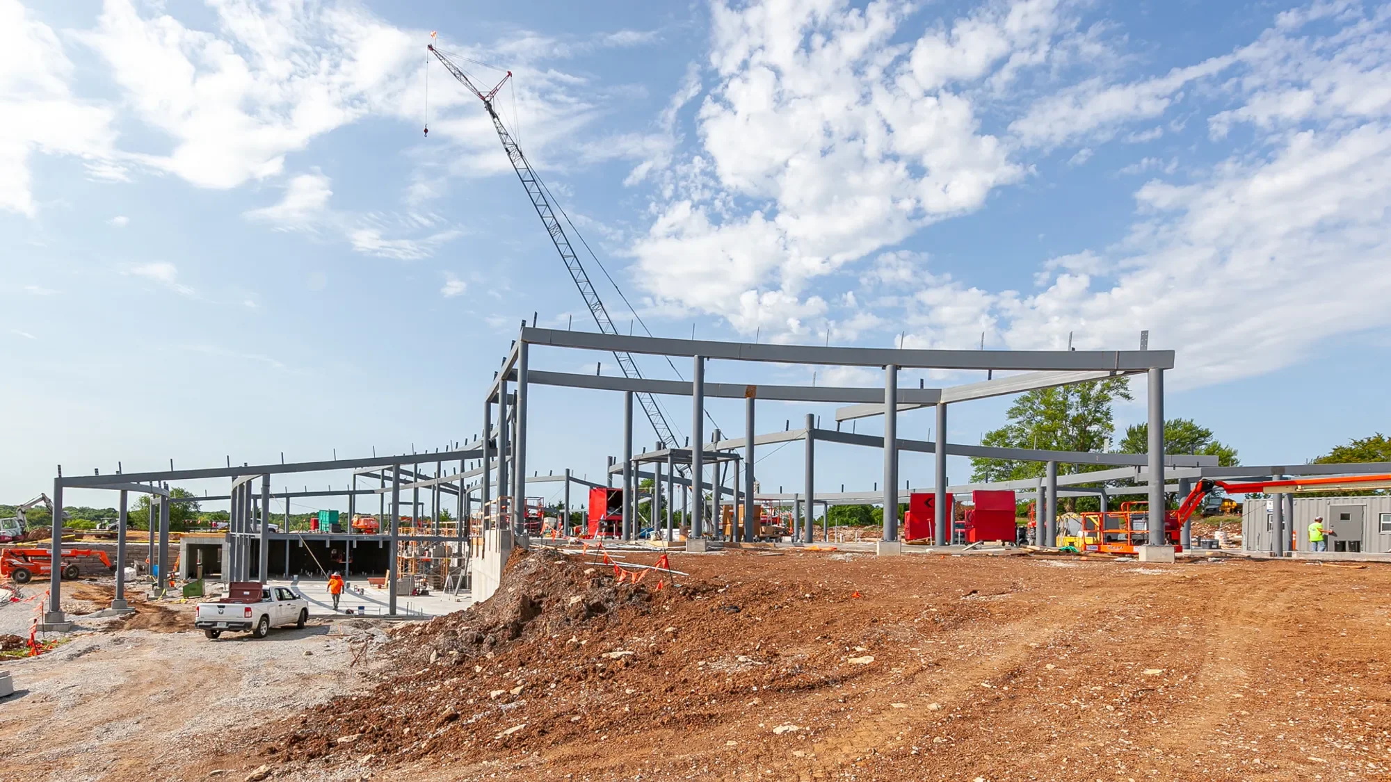 Construction site with metal framework structure, construction vehicles, and workers under a partly cloudy sky.