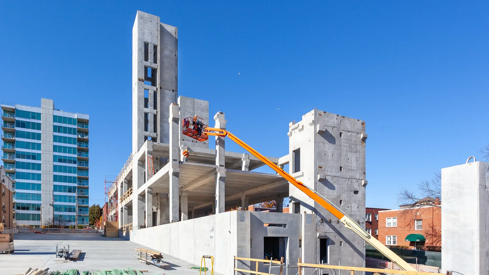 Construction site with partially built concrete structures, a worker operating a boom lift, and modern apartment buildings in the background under a clear blue sky.