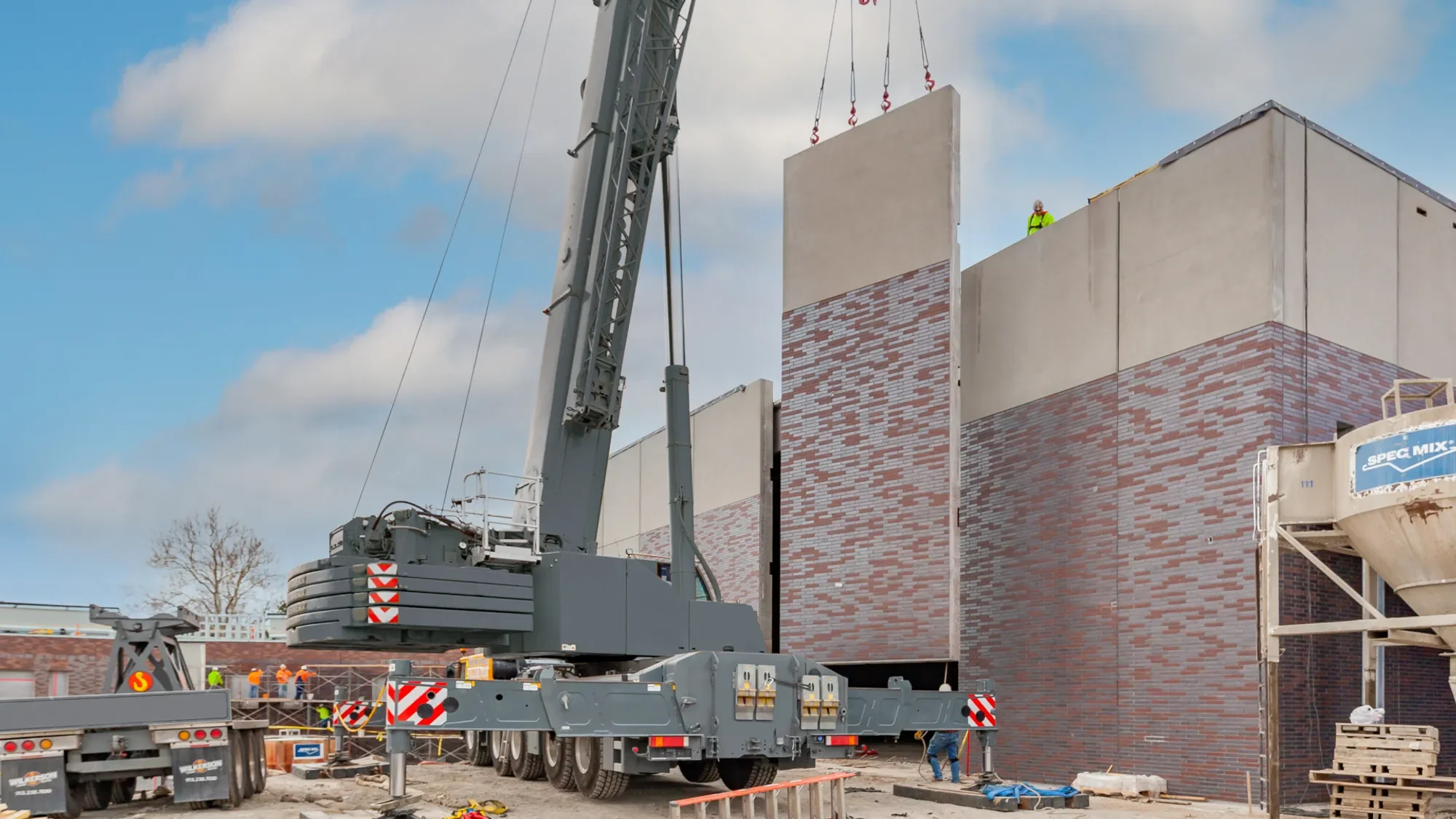 Construction workers operating a large crane lifting a wall section onto a building under construction. The building has red brick and concrete walls, with construction equipment and materials around the site.