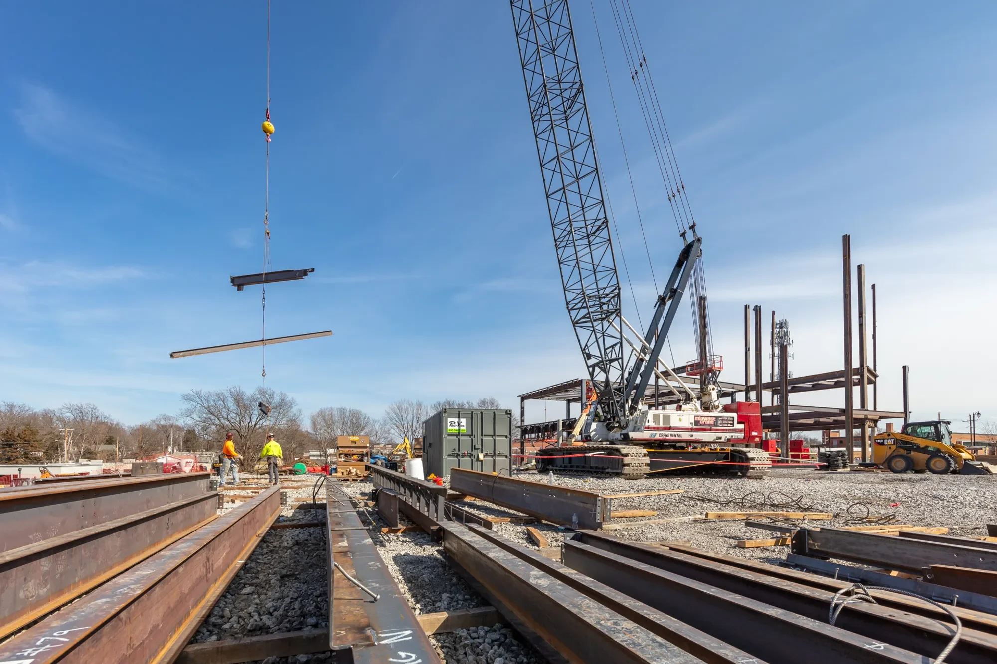 Construction site with steel beams being lifted by large crane. Two workers in safety vests and helmets are present. Steel beams are laid out on the ground, and various construction equipment and materials are visible. Clear blue sky in the background.