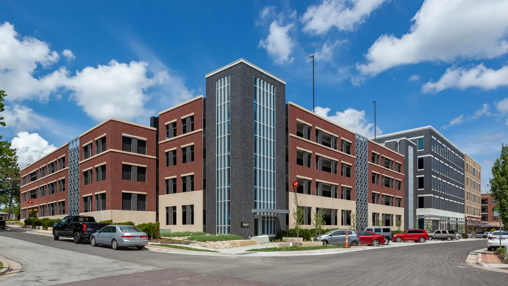 Modern multi-story building with red brick and black accents, large windows, and a parking lot in front under a partly cloudy sky.
