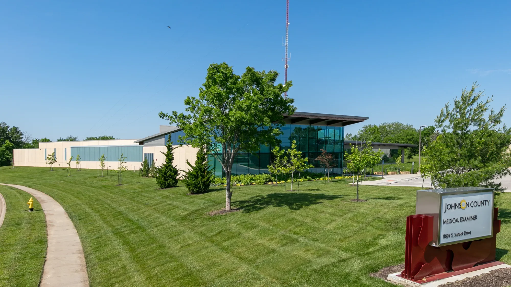 Modern medical office building with large glass facade, surrounded by well-manicured lawn and trees, under a clear blue sky; a sign in the foreground reads 'Johnson County Medical Examiner' with an address.