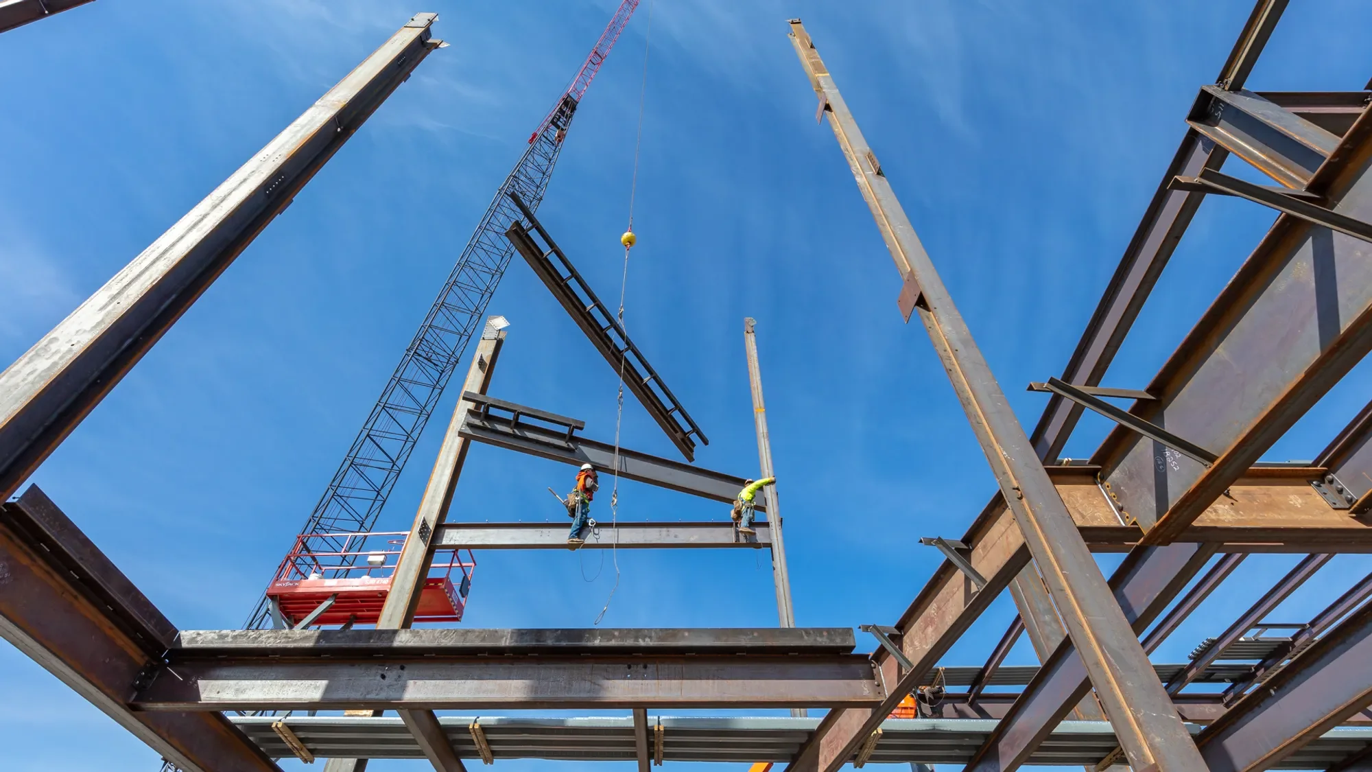 Construction workers on steel beams at a building site, with a crane lifting materials in the background, under a blue sky.