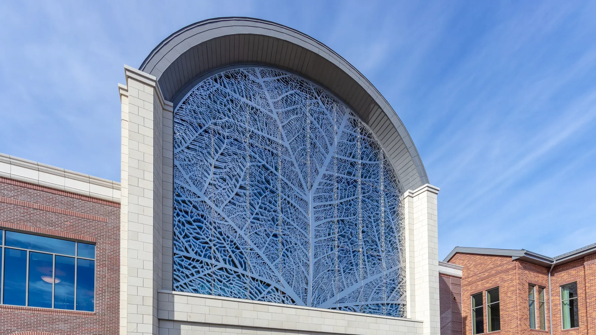 A brick and white stone building with a large arched window covered by a decorative metal screen resembling tree branches. Blue sky with some clouds in the background.