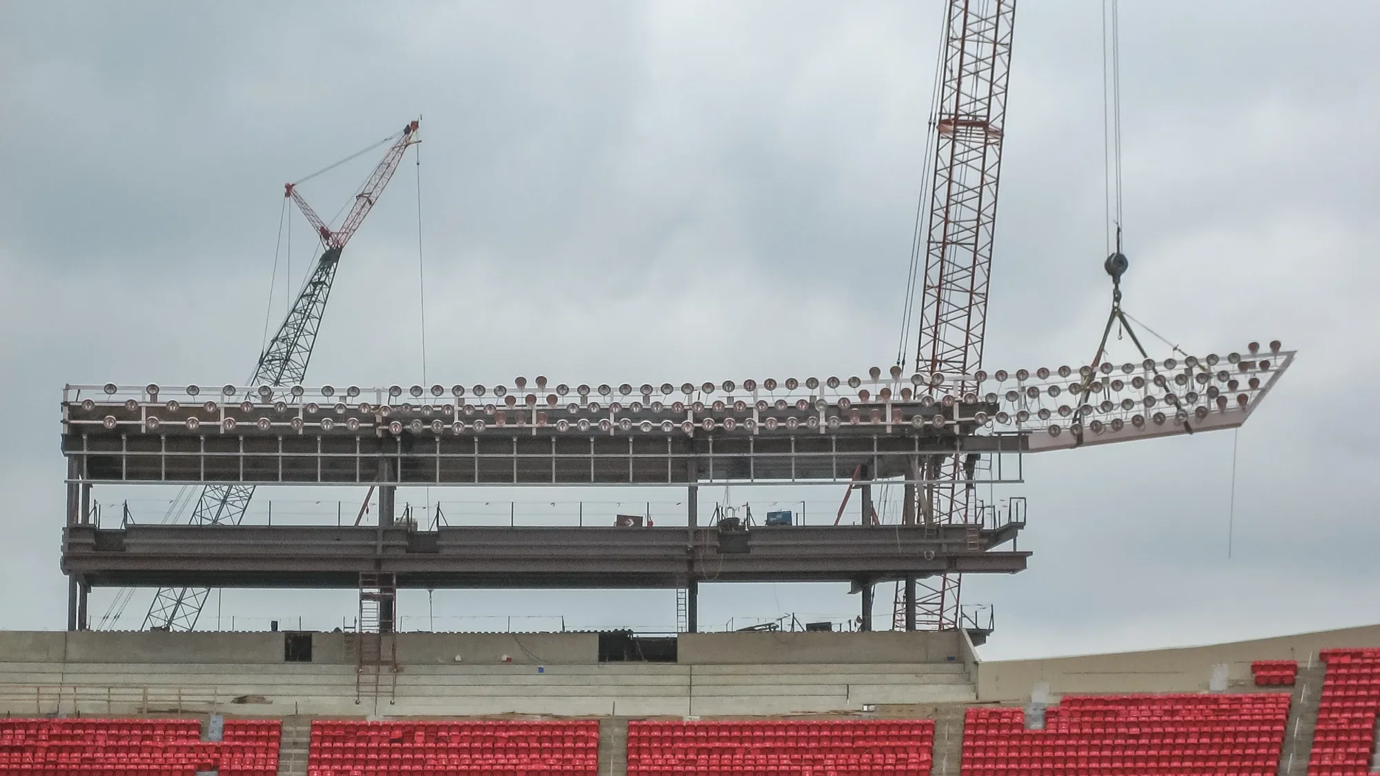 Construction site with a multi-level scaffolding structure, two tall cranes, and overcast sky in the background.